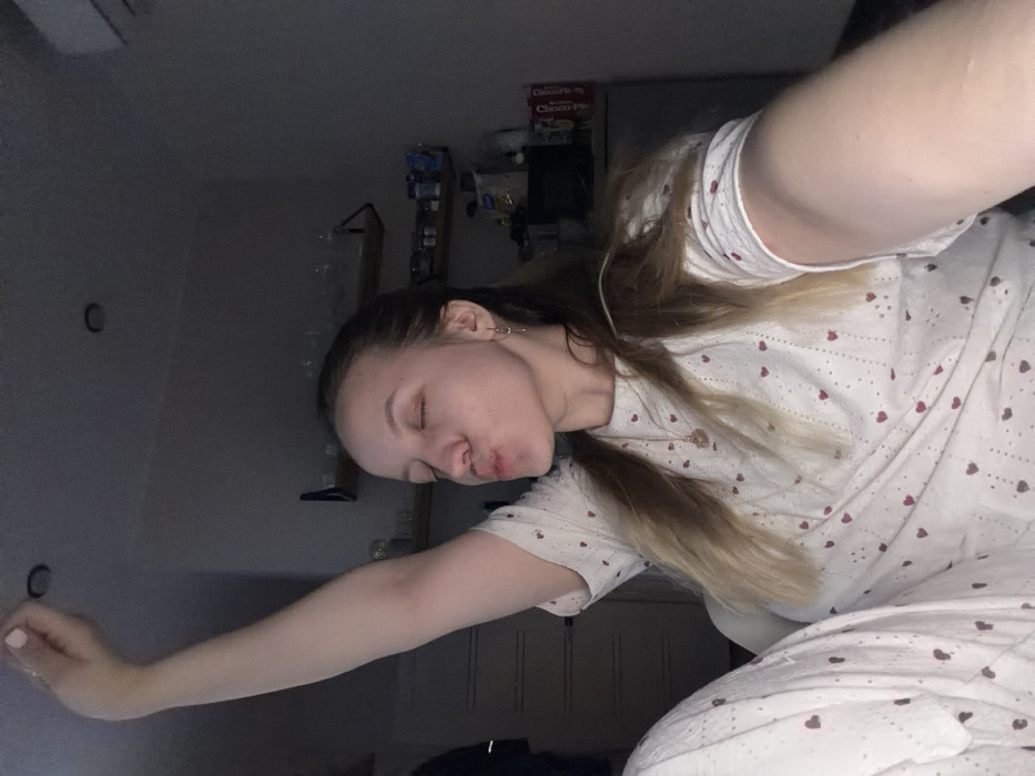 The scene depicts an interior setting, identified as being within the city of Shipulino, Russia. A young adult female with light skin and light brown hair, pulled back, is positioned centrally. Her eyes are closed, and her lips are pursed forward in a "kiss face" gesture. Her left arm is raised vertically above her head, with the hand curled into a fist, displaying light-colored fingernail polish. Her right arm, holding the camera, is extended forward and slightly to the right, out of frame. She is wearing a white short-sleeved top and matching pants, both patterned with small red hearts and vertical lines of tiny dots. A light-colored necklace with a small pendant and small hoop earrings are visible.
The background is dimly lit and appears to be a domestic space, potentially a kitchen or a combined living/kitchen area. To the left, a dark vertical object is partially visible. Centered in the background are two wooden shelves mounted on a light-colored wall, holding clear drinking glasses. Below these shelves, a counter or lower shelf area contains various containers and items, including a package with "CHOCO" visible on its label. A dark, possibly metallic, cabinet or refrigerator stands to the right. Two recessed circular light fixtures are visible in the white ceiling. The overall lighting suggests ambient or low artificial light.