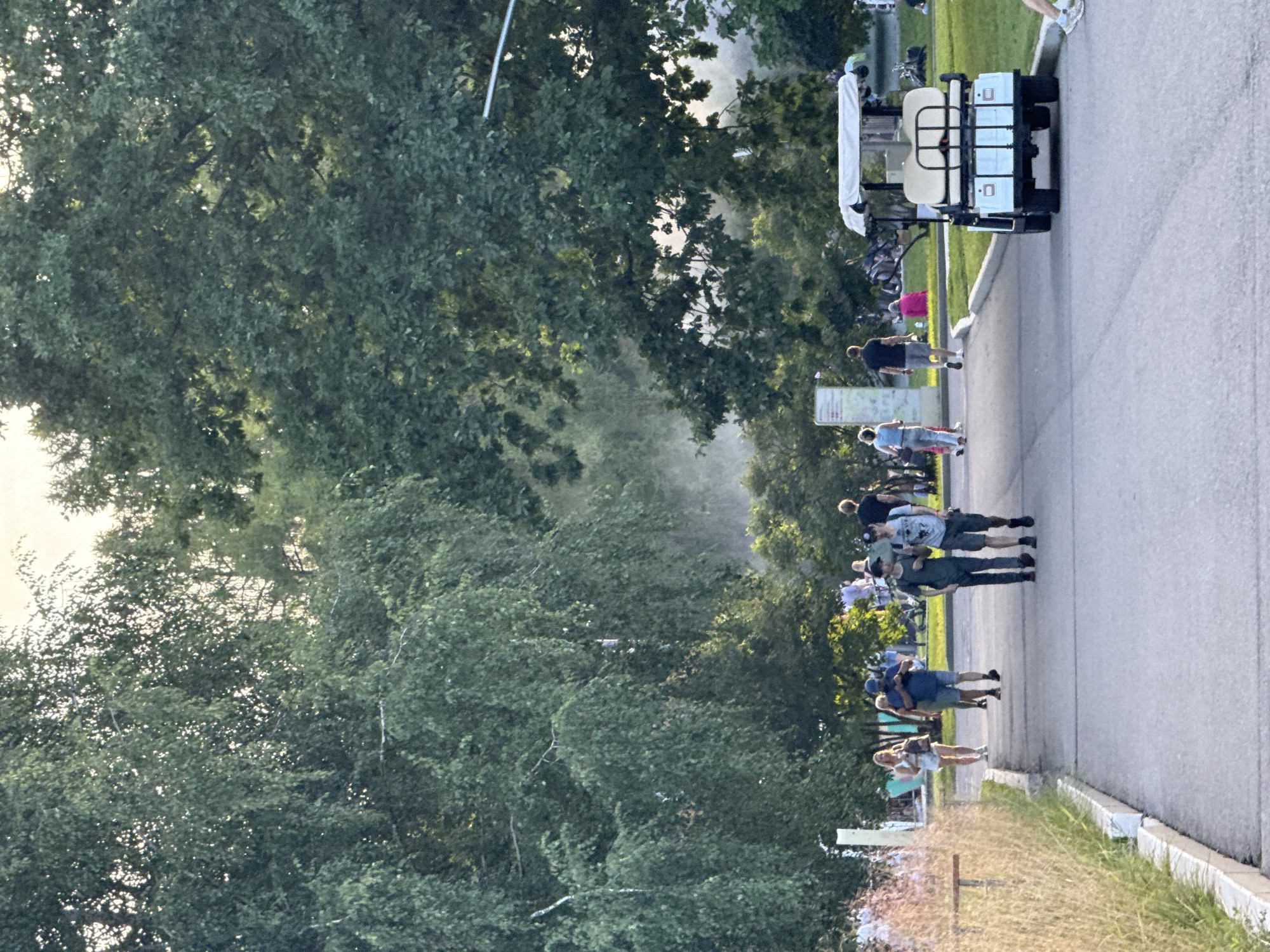 In Moscow, Russia, a paved pathway winds through a park with lush green trees and tall grass on the left. Several people are walking along the path. In the foreground, a white golf cart is parked on the right side of the path. In the background, more people are seen gathered on the grass, with some sitting and others standing. The overall scene depicts a relaxed atmosphere in a park setting during daylight hours, with a slightly hazy quality to the air in the middle ground.