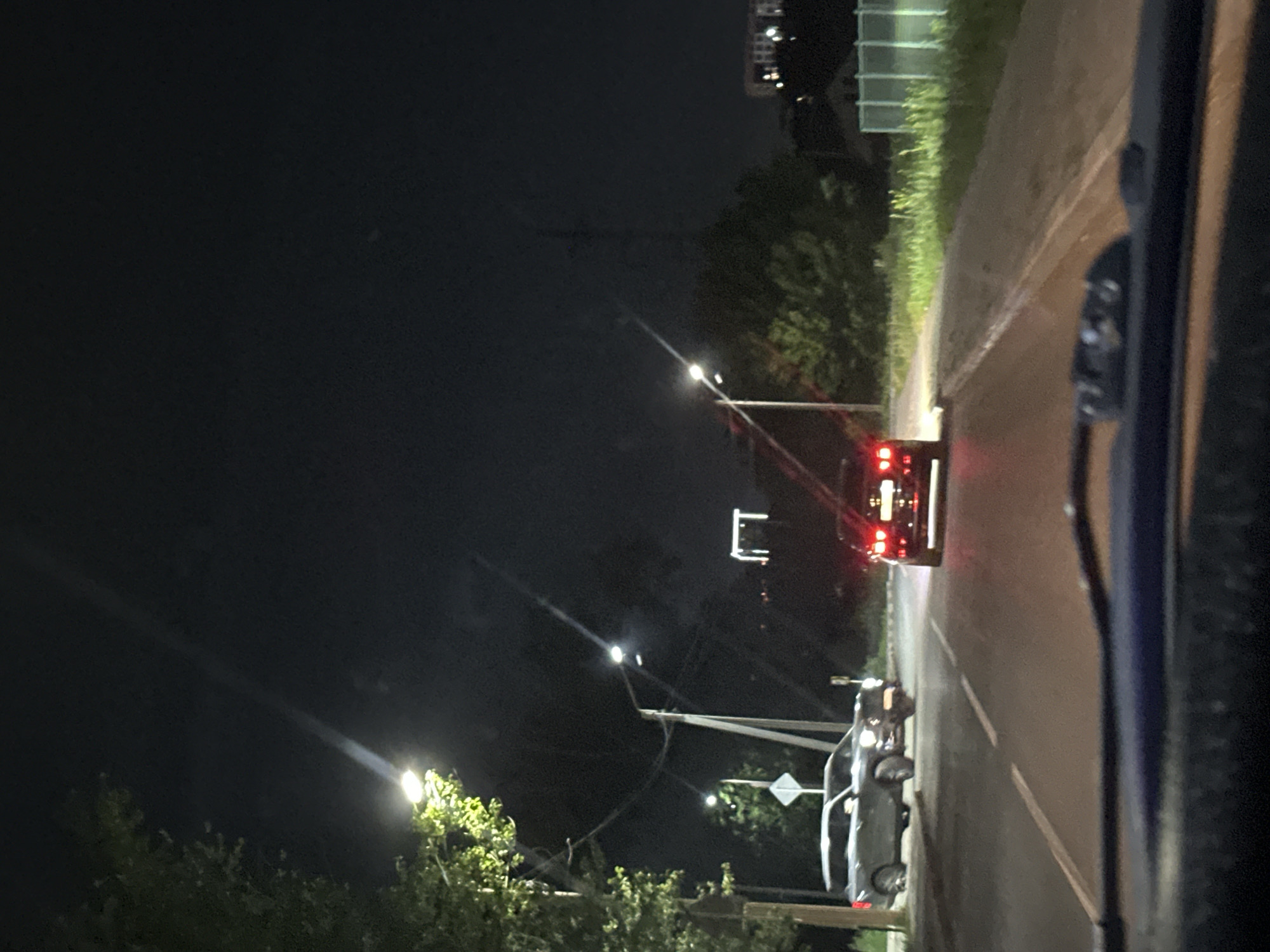 The image depicts a nighttime street scene, likely within the city of Kostanay, Kazakhstan. The perspective is from inside a vehicle, looking forward through the windshield. The road is paved and marked with white lane lines.
In the foreground, the top portion of the vehicle's dashboard is visible, out of focus.
On the road, two cars are visible. A darker colored SUV is in the left lane, facing away from the camera. To its right and further down the road, a darker sedan is also visible, facing away from the camera, its taillights illuminated.
Streetlights are present, casting pools of light on the road and illuminating the surrounding vegetation and trees. One streetlight is prominent on the left side of the image, with a visible beam of light. Another streetlight is visible further down the road on the right.
In the background, trees and vegetation line the sides of the road. Buildings and structures are faintly visible in the distance, suggesting an urban or suburban environment. A tall, rectangular structure, possibly a building or pylon, is illuminated by lights in the middle distance. A fence, possibly a green chain-link fence, is visible on the right side of the road in the background. The sky is dark, indicating nighttime.