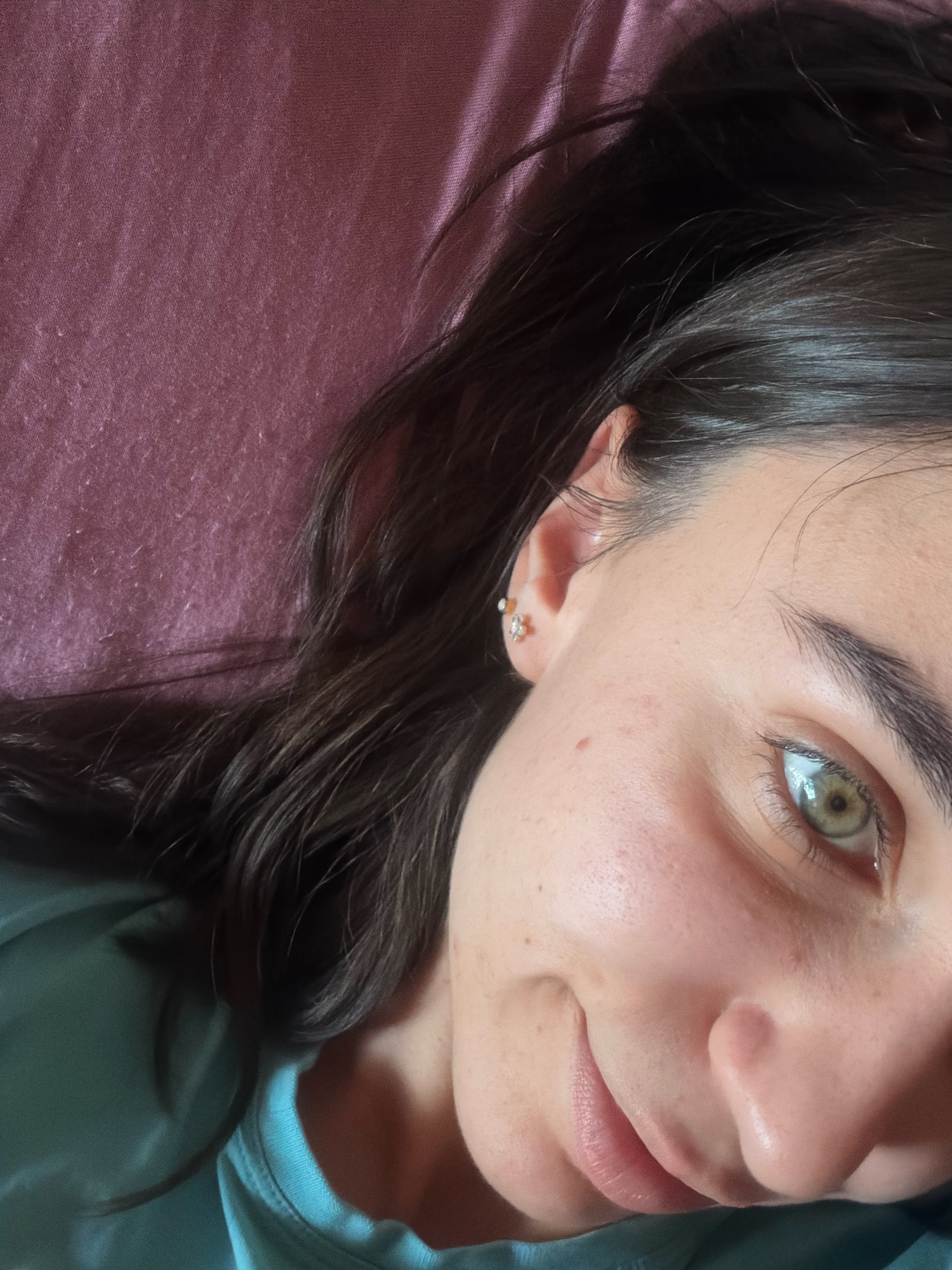 The image shows a close-up, top-down view of a young woman's face as she lies on a textured, plum-colored surface. Her dark hair is spread out around her head. She is wearing a teal-colored t-shirt. The woman's left eye, which is a striking green, is in focus, along with part of her nose and lips. She has a faint smile on her face and a small mole on her left cheek. She is also wearing a small, star-shaped earring in her left ear. The lighting suggests an indoor setting, possibly a bedroom. The image does not provide any specific information about the location in the city of Sofia, Bulgaria.