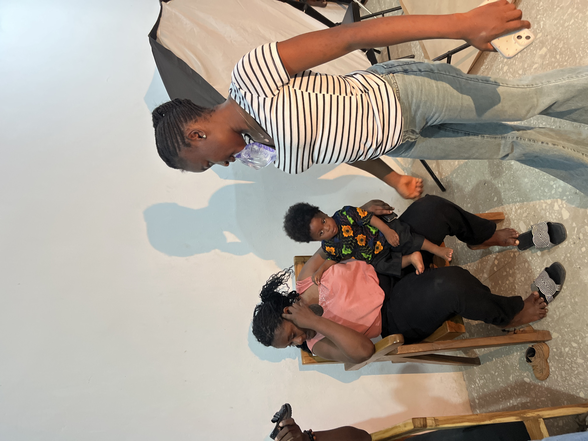 A group of people, including a woman holding a baby, are in what appears to be a studio setting in Gura Top, Nigeria. The woman holding the baby is seated in a wooden chair, wearing a pink top and black pants. The baby is dressed in a colorful, patterned shirt. Another woman, wearing a striped shirt and jeans, is standing and appears to be interacting with the seated woman. A softbox light is visible in the background, suggesting a professional photo or video shoot. The environment is simple, with a white wall as a backdrop. The floor is tiled.