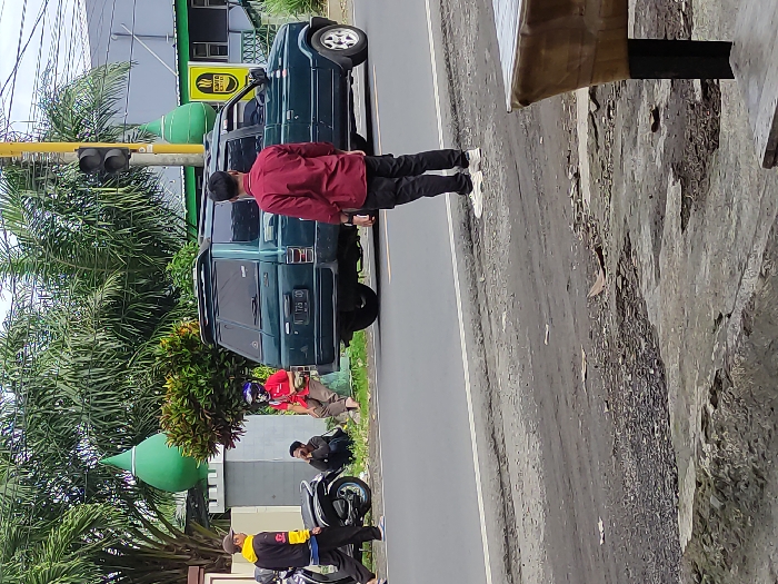 This outdoor street scene in Kepanjen, Indonesia, depicts a roadside environment on what appears to be an overcast day. The image is taken from an elevated perspective, showcasing a paved road with a white demarcation line on the right, and a dirt shoulder leading to an area with buildings, trees, and people on the left.

The central figure is a man in a maroon long-sleeved shirt, black pants, and white sneakers, standing with his back to the camera. He is positioned next to a dark green SUV, which resembles an older model, possibly a Toyota Kijang. The SUV bears an Indonesian license plate, "N 725 GD."

Behind the SUV, a black traffic light pole is visible. Further down the street, several other individuals are present: a person in a red shirt and a helmet is seated or crouching, and two more people are seen sitting on a low wall or the ground. In the lower-left foreground, a man wearing a yellow and black jacket stands beside a black motorcycle or scooter.

The background is rich with tropical foliage, including numerous palm trees and various bushes. A distinct green dome-shaped structure, possibly part of a mosque, is prominent among the greenery. Adjacent to it is a green building with a yellow sign that clearly displays the word "KOPI" (coffee), suggesting a coffee shop or cafe. On the far right, a partial view of a wooden or canvas structure can be seen. The overall scene captures a calm, everyday street activity.