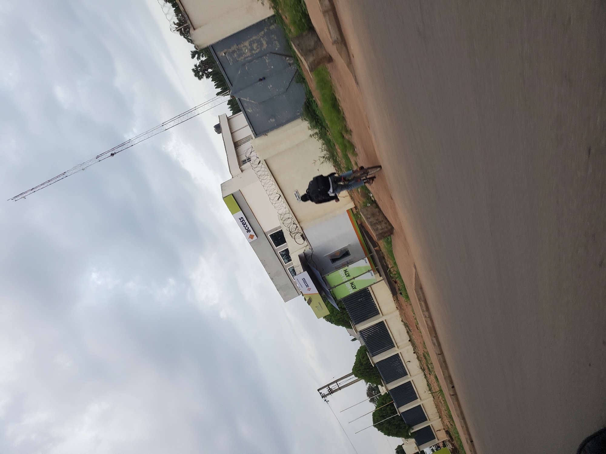 In Bukuru, Nigeria, on a cloudy day, a person rides a bicycle along a road adjacent to a white building with a gray gate and barbed wire on top of the fence. The building has the "Access" logo and "24HR ATM" signage. A tall radio tower stands in the background. The road is paved and slopes downwards from right to left, with a concrete sidewalk and grass bordering the building.