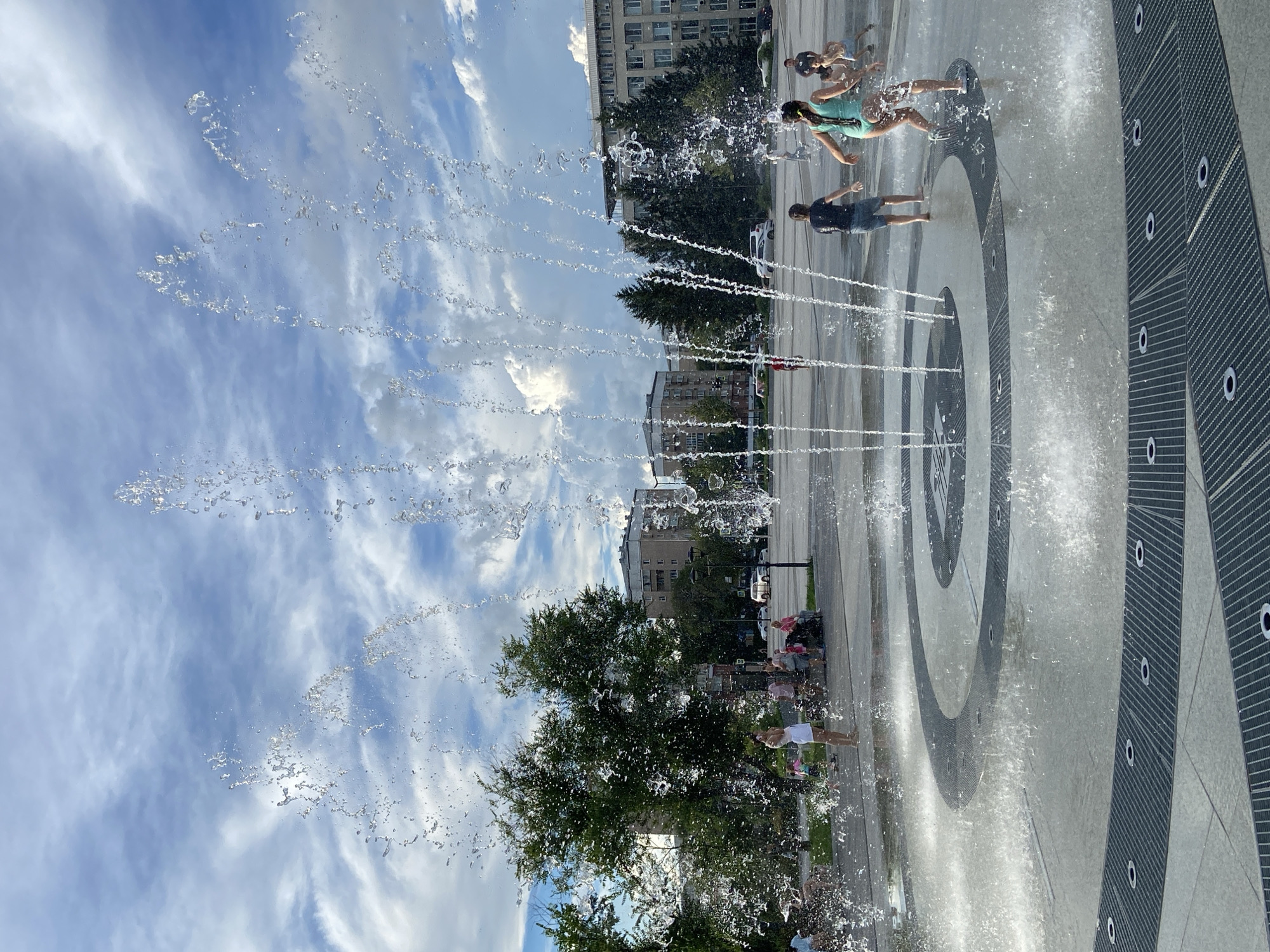 In Blagoveshchensk, Russia, children and adults are enjoying a warm day at a public fountain. The fountain is located in an open, paved area with concentric circles on the ground. Water sprays upwards in multiple streams, creating a refreshing spectacle. Several children are actively playing in the water, their movements captured mid-splash. Nearby, adults observe from the side, some seated and others standing. In the background, lush green trees stand tall against a backdrop of multi-story buildings. The sky is a vibrant blue, dotted with fluffy white clouds, suggesting pleasant weather. The scene captures a moment of leisure and recreation in an urban setting.