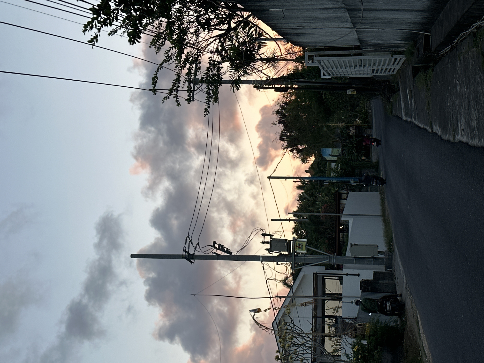 This media file captures a street scene in Kuta Selatan, Indonesia, during what appears to be dusk. The sky is filled with dramatic, multi-colored clouds, hinting at the sunset, with hues of pink, orange, and grey dominating the scene. Overhead, a complex network of power lines crisscrosses the frame, supported by utility poles. These lines are partially obscured by the branches of trees laden with leaves.
On the right side of the image, a paved road runs alongside a concrete wall and a white fence. The road is mostly empty, with a single person on a scooter visible in the distance, heading away from the viewer. Further down the road, another motorcycle is parked next to a building.
The buildings in the foreground are simple, with white walls and dark-framed windows. One building has an external electrical transformer mounted on a pole. The vegetation in the scene is lush, with trees and plants adding a tropical feel to the environment. The overall mood is serene, characteristic of twilight in a tropical locale. No visible text is discernible in the image.