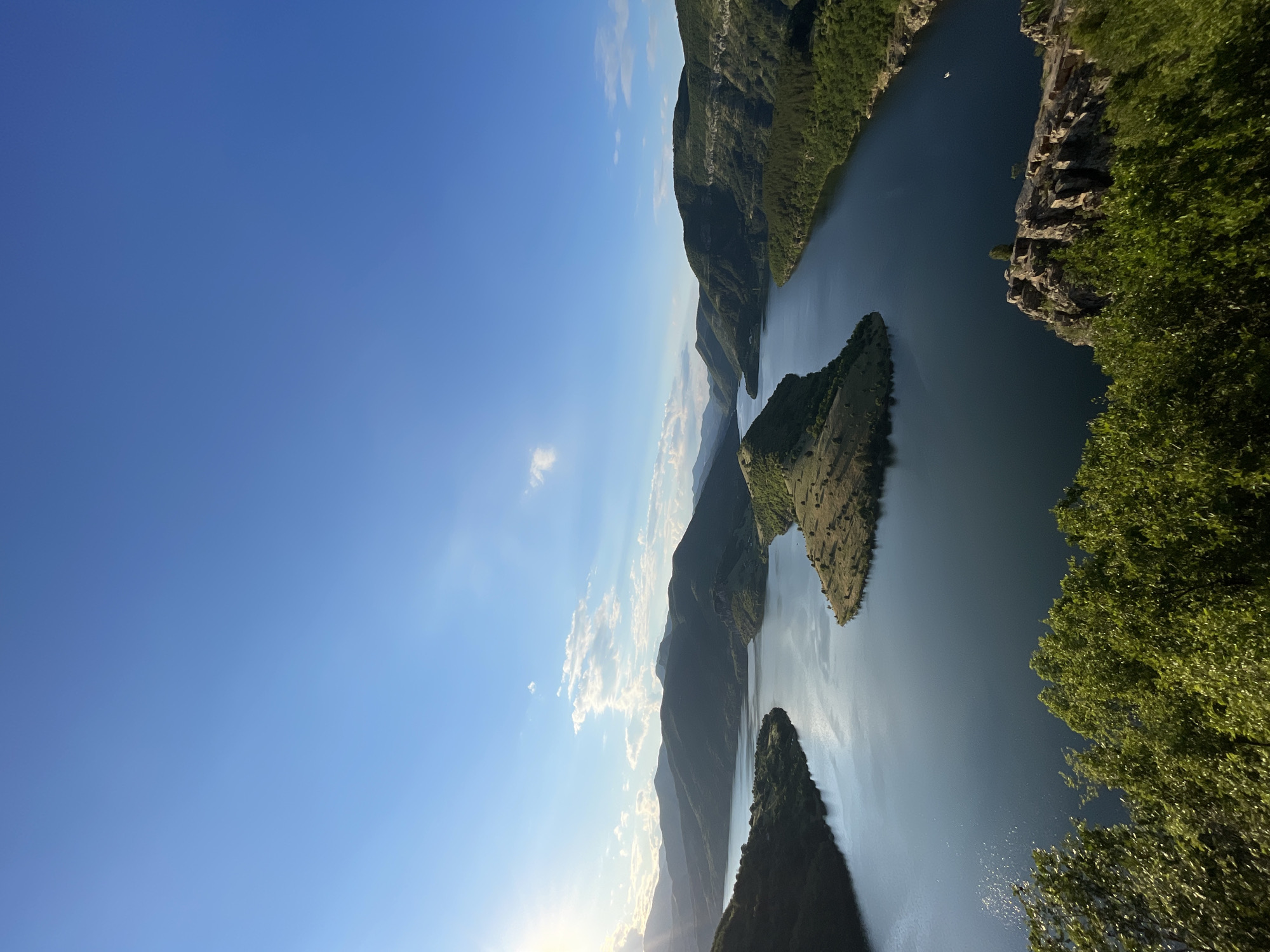A panoramic view captures the Arda River in Borovitsa, Bulgaria, under a clear blue sky dotted with sparse white clouds. The sun, positioned in the upper left quadrant, casts a soft glow, illuminating the scene. A prominent, heart-shaped island covered in dry grass sits in the center of the expansive river, surrounded by steep, verdant hills. The water's surface reflects the sky and the surrounding landscape, creating a serene and tranquil atmosphere. Lush green trees frame the foreground, adding depth and texture to the composition. A small boat can be seen on the right side of the river, hinting at human presence in this natural landscape. The overall scene evokes a sense of peace and natural beauty.