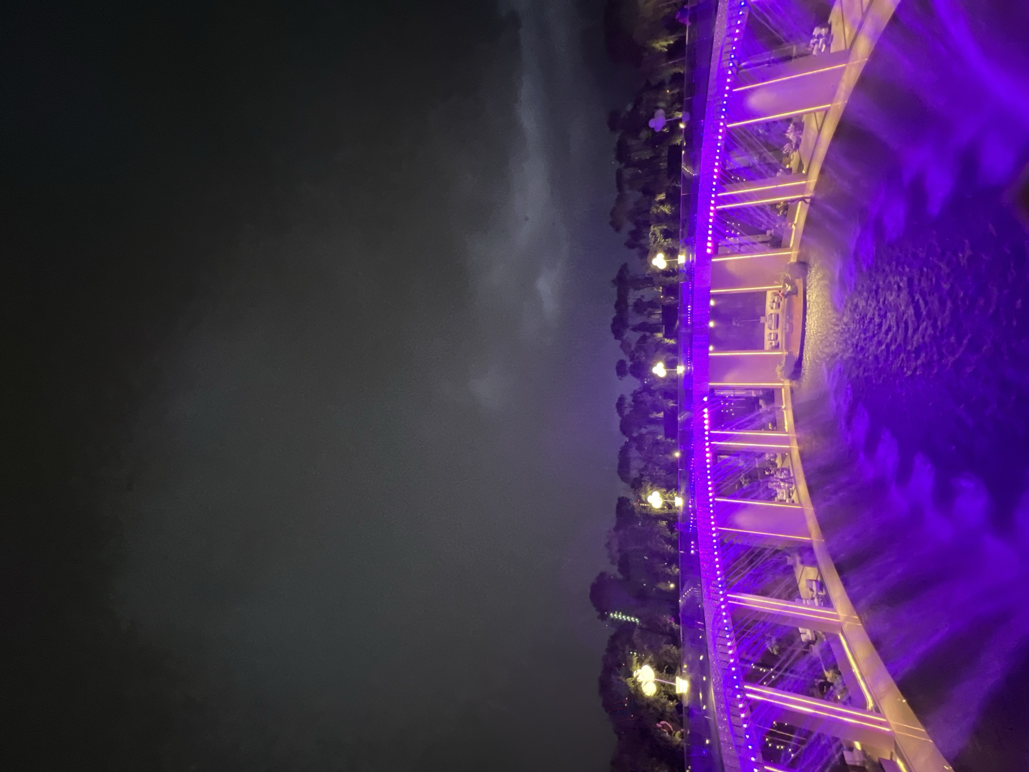 The image depicts a nighttime scene at the Oasis Aqua Park in Krasnodar, Russia. A contemporary, curved bridge illuminated with vibrant purple lights spans over a body of water. The water itself is also illuminated with purple light, creating a luminous effect. Tables and seating are visible on the bridge, suggesting it is part of a dining or lounge area. A backdrop of dark trees and a cloudy, dark sky frames the scene.