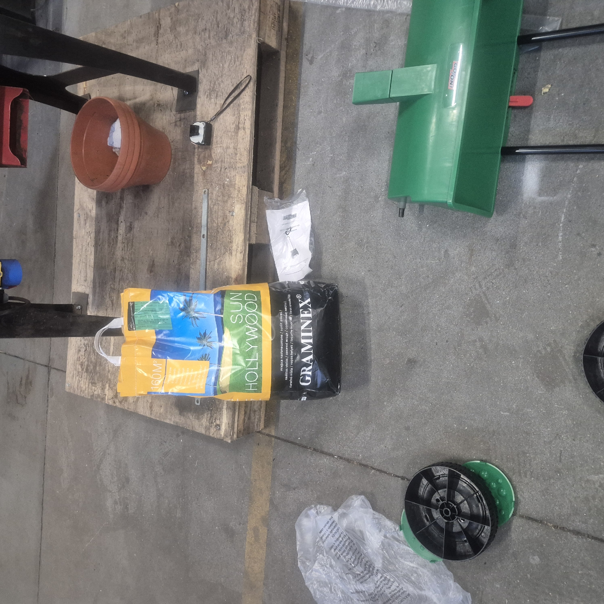 The image depicts a scene on a concrete floor, possibly in a storage area or workshop, in Nowa Wieś Książęca, Poland. In the center of the image, there is a large yellow and blue bag of grass seed labeled "SUN HOLLYWOOD" and "GRAMINEX". The bag is positioned on a wooden pallet. To the right of the bag, there is an orange clay pot containing some white material. A small black and white measuring tape is also visible on the pallet. In the lower left corner, a black wheel with green accents is placed on the ground. To the right and slightly further back, a green gardening tool with black legs is visible. A crumpled plastic bag is partially visible to the left of the seed bag. The environment is utilitarian, with a concrete floor and industrial elements like a metal frame supporting some equipment in the background. There are no people visible in the image.