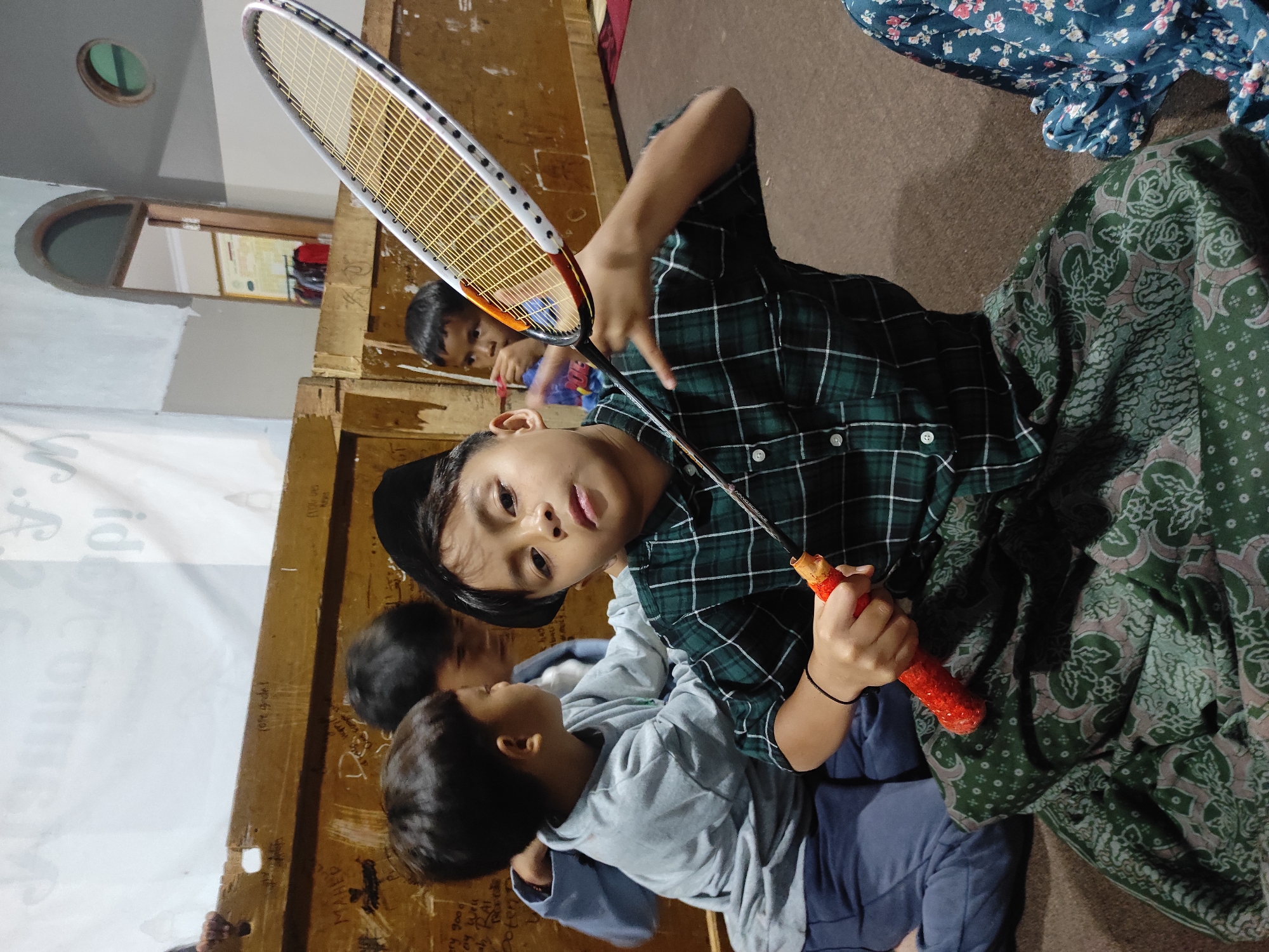 In Sumedang, Indonesia, four young boys are gathered indoors, with one in the foreground holding a badminton racket. The boy in the foreground, wearing a dark green plaid shirt and a black cap, is seated and looking towards the camera, with the racket held out in front of him. He is also wearing a patterned sarong. Behind him, three other boys are visible. One boy in a light gray shirt and dark pants is seated to the left. Another boy, wearing a blue shirt, stands behind and to the left of the boy with the racket. The fourth boy, wearing a blue shirt, is partially visible on the right. The environment appears to be a room with wooden walls and a patterned carpeted floor. The background shows a doorway with a framed poster and some hanging clothes.