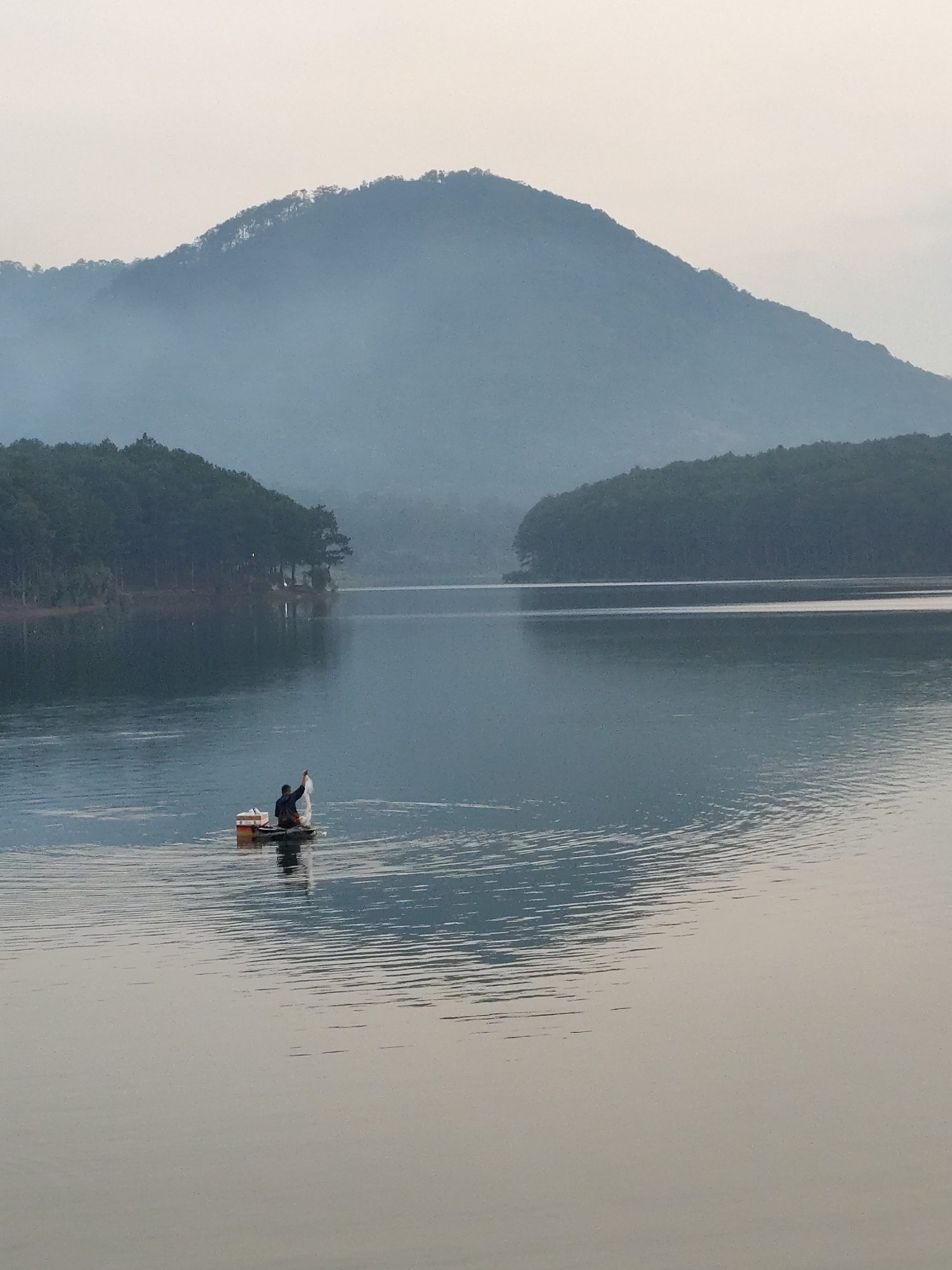 The image depicts a tranquil lake scene in Da Lat, Vietnam. A lone fisherman is visible in the center, standing on a small raft, holding what appears to be a fishing net above the water. The calm water reflects the surrounding landscape, including a large, misty mountain in the background and forested hills on either side. The sky is overcast, creating a soft, muted light over the entire scene.