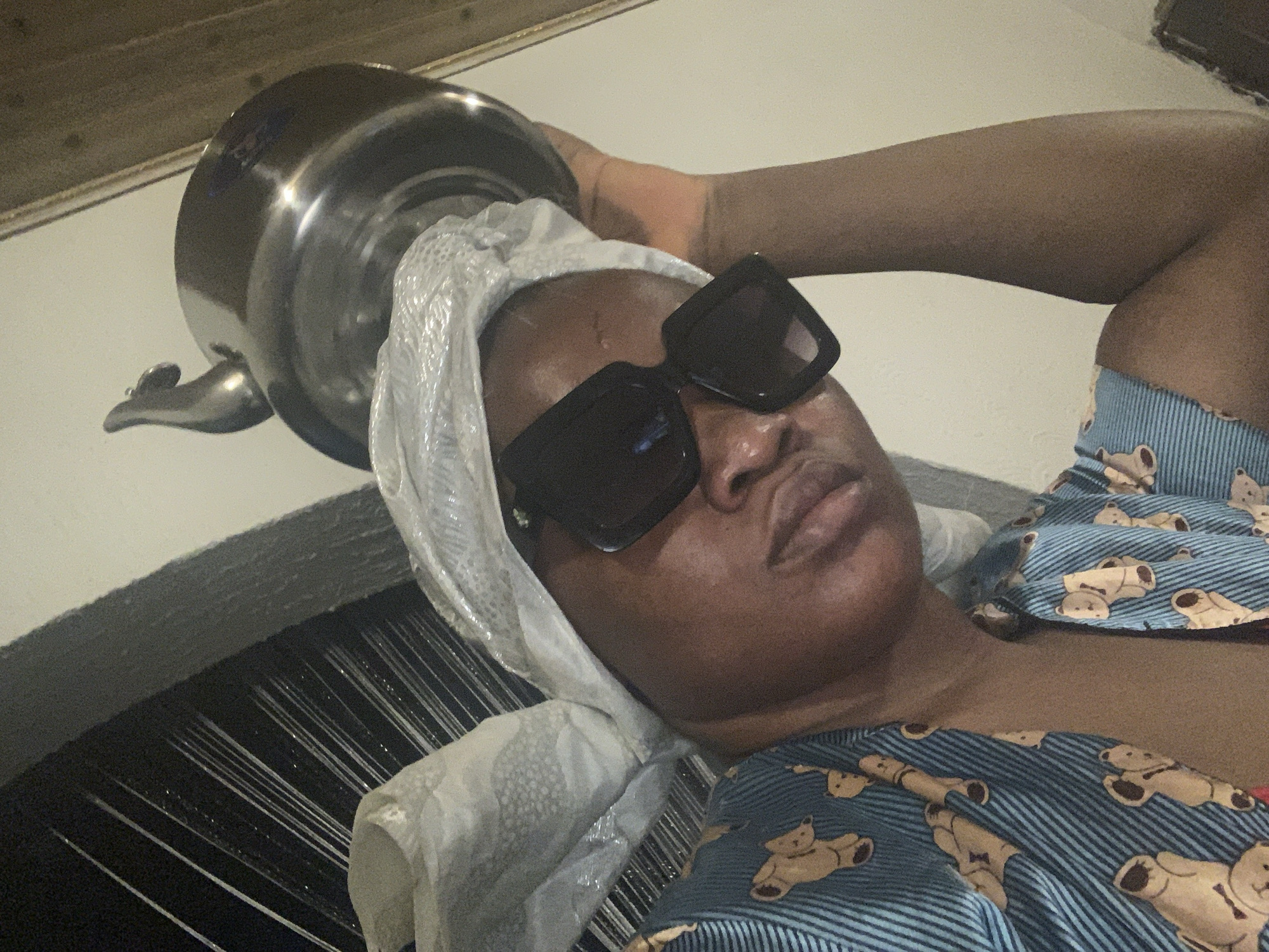 A person wearing sunglasses and a plastic head covering is lying in a bed with a large metal pot resting on their head. The bed has a black headboard with linear designs. The person is wearing a blue and white striped shirt with teddy bear patterns. The location is inferred to be Barakin Akawo, Nigeria.