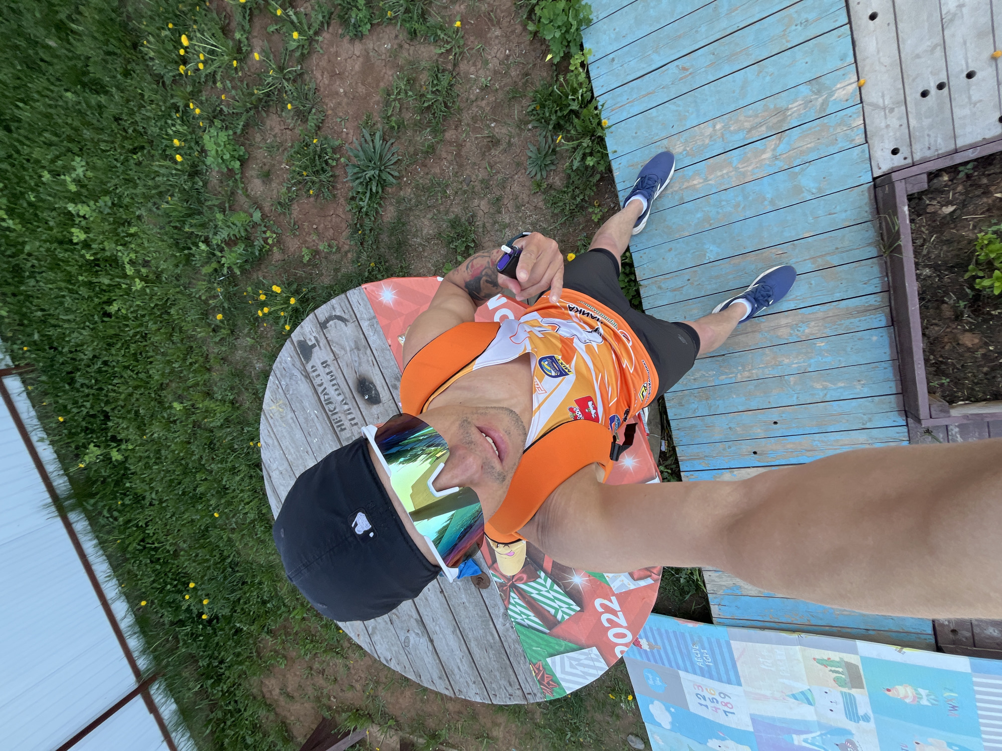 A man is taking a selfie from a high angle, standing on a large, round, wooden spool with painted holiday decorations. The man is wearing a black baseball cap, reflective sunglasses, an orange and white athletic shirt, and black shorts. He has a tattoo on his left arm and is holding a small black object in his left hand. He has blue sneakers on. He is standing on a wooden spool, which is on a patch of grass and dirt, partially covered in yellow flowers. Behind him is a walkway made of blue wooden planks and a small garden plot. The location appears to be in Chaikovsky, Russia, based on the Cyrillic writing on the spool. A white metal structure is visible on the left side of the frame.