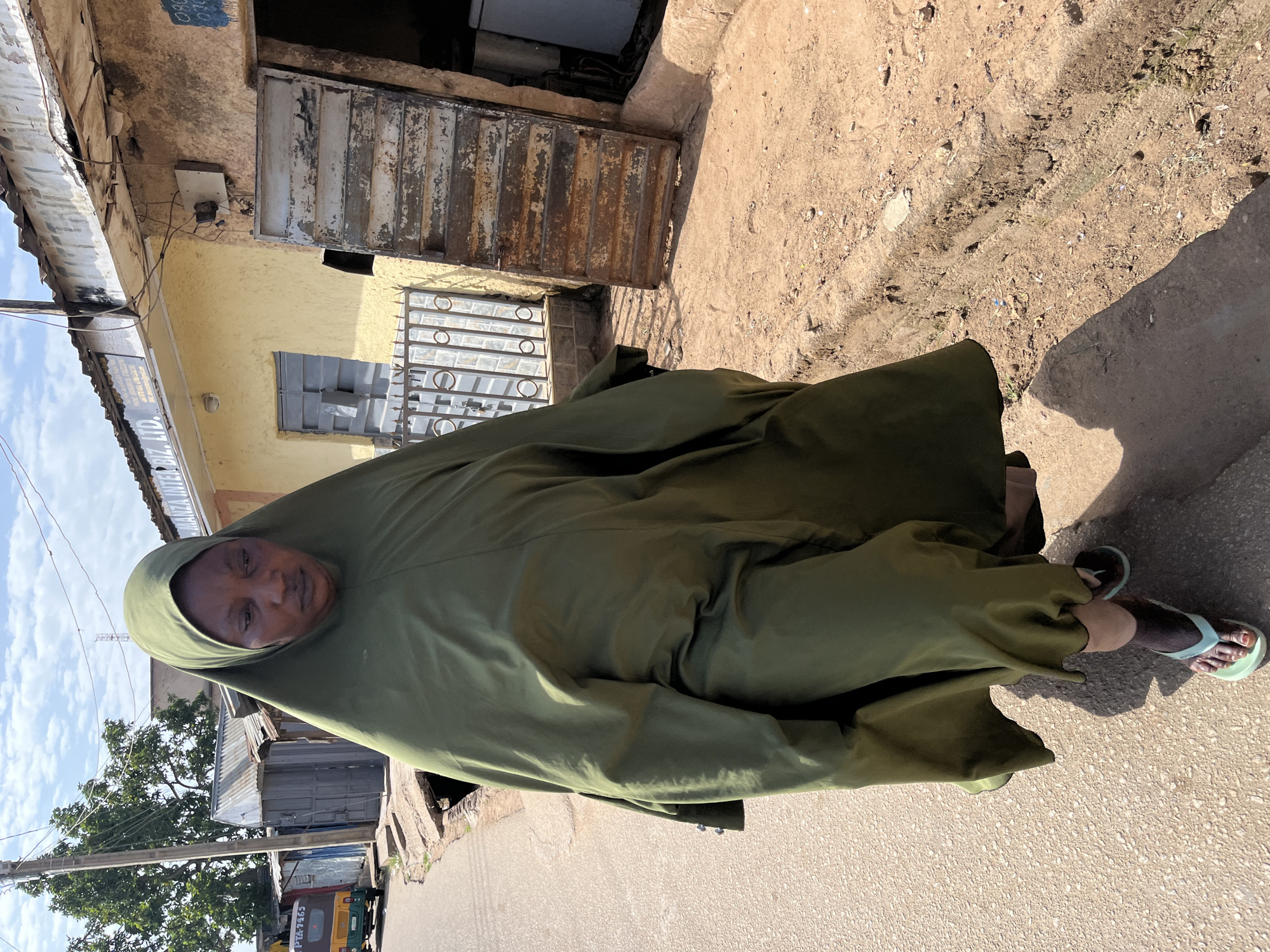 A woman wearing a green hijab stands on a street in Jos, Nigeria. The street is paved and partly shaded. Buildings are visible, including one with a yellow facade and a rusty metal door. A vehicle is partially visible on the left side of the image. The woman is looking directly at the camera. She is wearing brown sandals with a light blue strap.