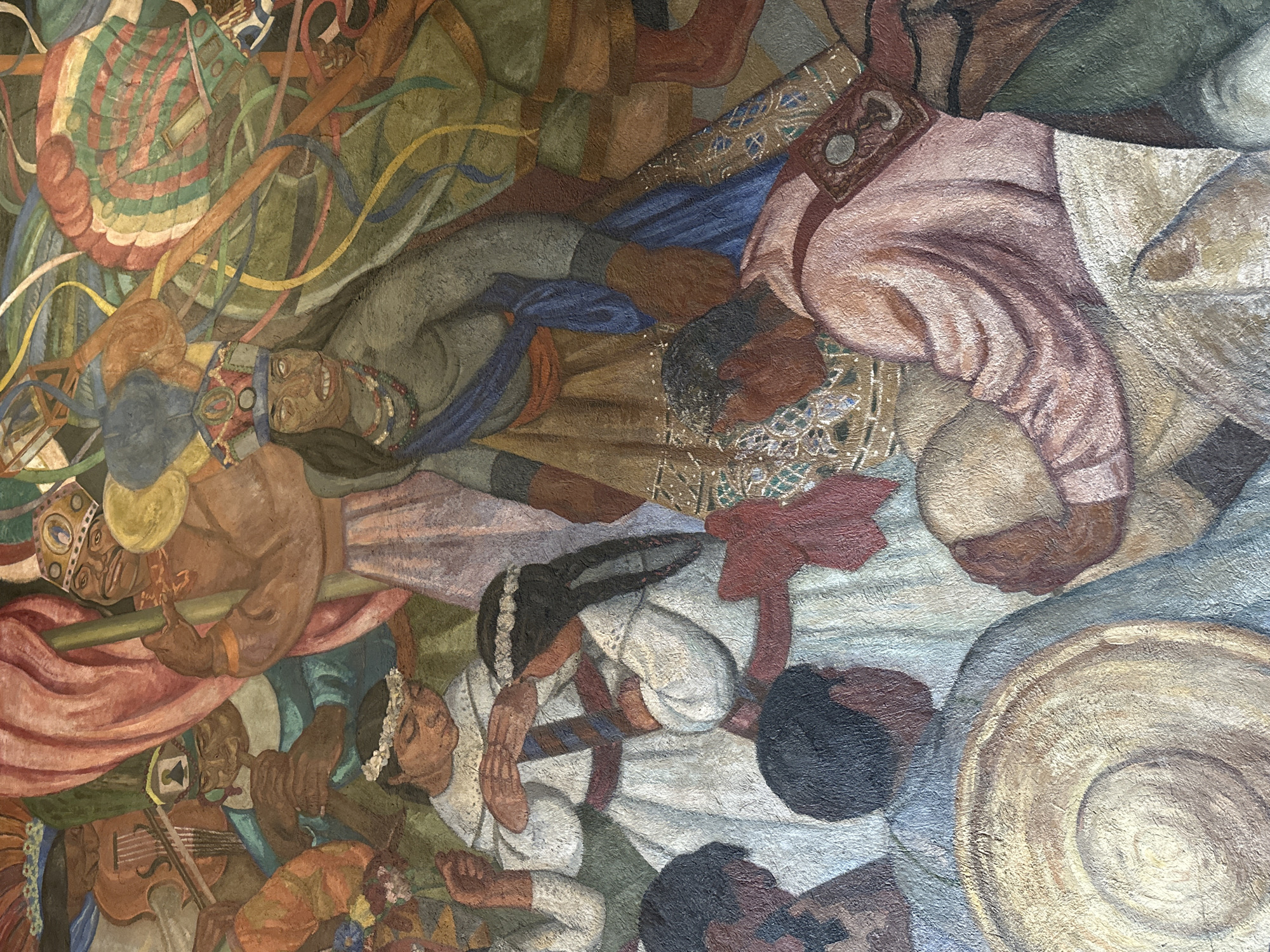 The image shows a section of a mural in Mexico City, Mexico. It depicts a chaotic scene with multiple figures. A figure with a large headdress and exposed teeth is central, surrounded by colorful ribbons and objects. Below, figures are playing instruments, including a violin, and others are resting. The overall composition is dense and crowded, with a mix of people and objects rendered in earthy tones. The style suggests a public art mural.