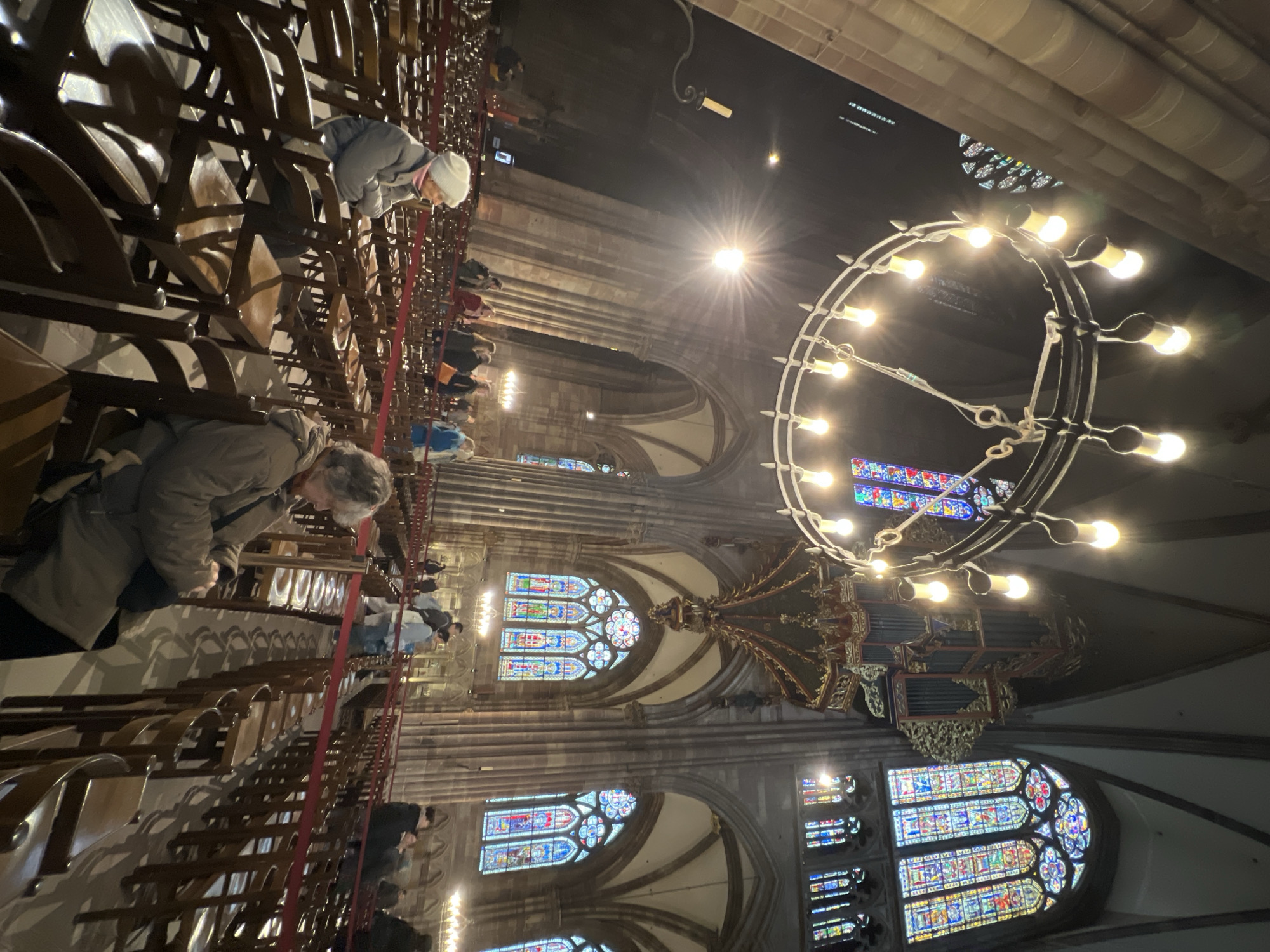 The image captures the interior of the Strasbourg Cathedral in Strasbourg, France. The focus is on a large, circular chandelier with multiple light bulbs, suspended from the ceiling. Below the chandelier, a view through arched windows filled with stained glass is visible.
The foreground contains rows of wooden pews. A number of people are seated, primarily women, some in prayer. A red rope barrier runs along the pews. The lighting is a mix of natural light filtering through the stained-glass windows and the artificial light from the chandelier. The architectural design is Gothic, with pointed arches and ribbed vaults typical of the style.