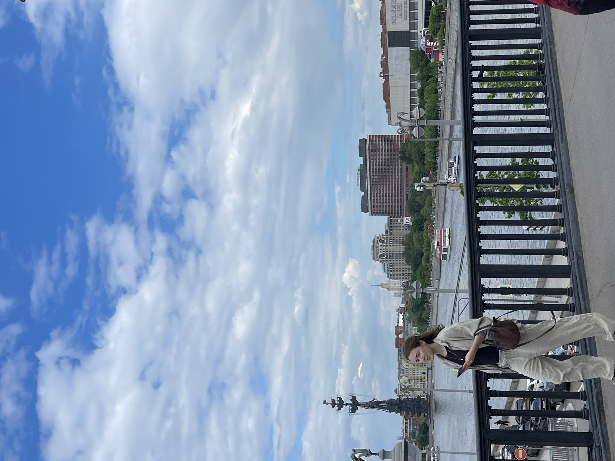 A woman stands on a bridge looking at a phone. The bridge has a black railing. In the background is a river and a city skyline, including multiple buildings and a statue of Peter the Great on the river, placing the location in Moscow, Russia. The sky is blue with scattered clouds.