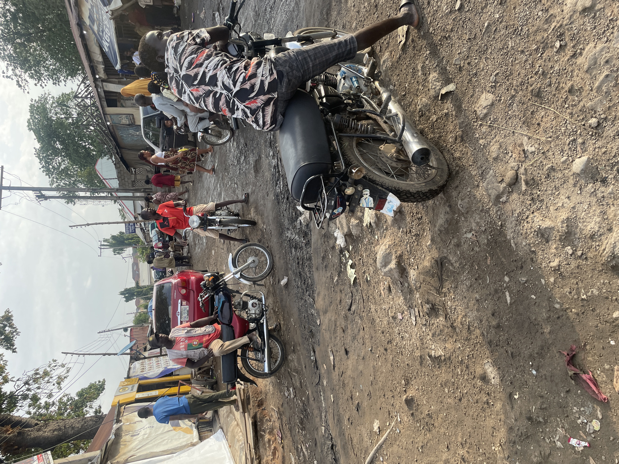 This image captures a vibrant, everyday street scene in Abuja, Nigeria, during daylight hours. The setting features a wide, unpaved dirt road, heavily trafficked by both pedestrians and motorcycles. On the left side of the street, various simple commercial buildings or residences with corrugated metal roofs line the thoroughfare, partially obscured by trees and overhead utility lines.
Numerous people are present, indicating a busy urban environment. In the immediate foreground, a man wearing a black, white, and red patterned shirt and plaid shorts is seated on a motorcycle, looking to his right. Another motorcycle is positioned further back. A red car, identifiable as a Toyota Corolla, is visible mid-ground, with a partially legible Nigerian license plate displaying "FEDERAL REPUBLIC OF NIGERIA" and "RC-57AE Abuja," confirming the location. A man in a red T-shirt, possibly a sports jersey with "PORTU" visible, sits on a motorcycle next to the car. Other individuals, including a woman in a patterned dress and various men, are walking or standing along the street.
The ground is a mix of dirt and small stones, with scattered litter such as plastic wrappers and sticks. The sky is bright but appears slightly hazy. The overall impression is one of active daily life in a bustling local area, with motorcycles serving as a primary mode of transportation. There are no strong discernible emotions among the individuals; they appear to be going about their routines. Visible text includes "RC-57AE Abuja" on the car's license plate and "PORTU" on a man's shirt.