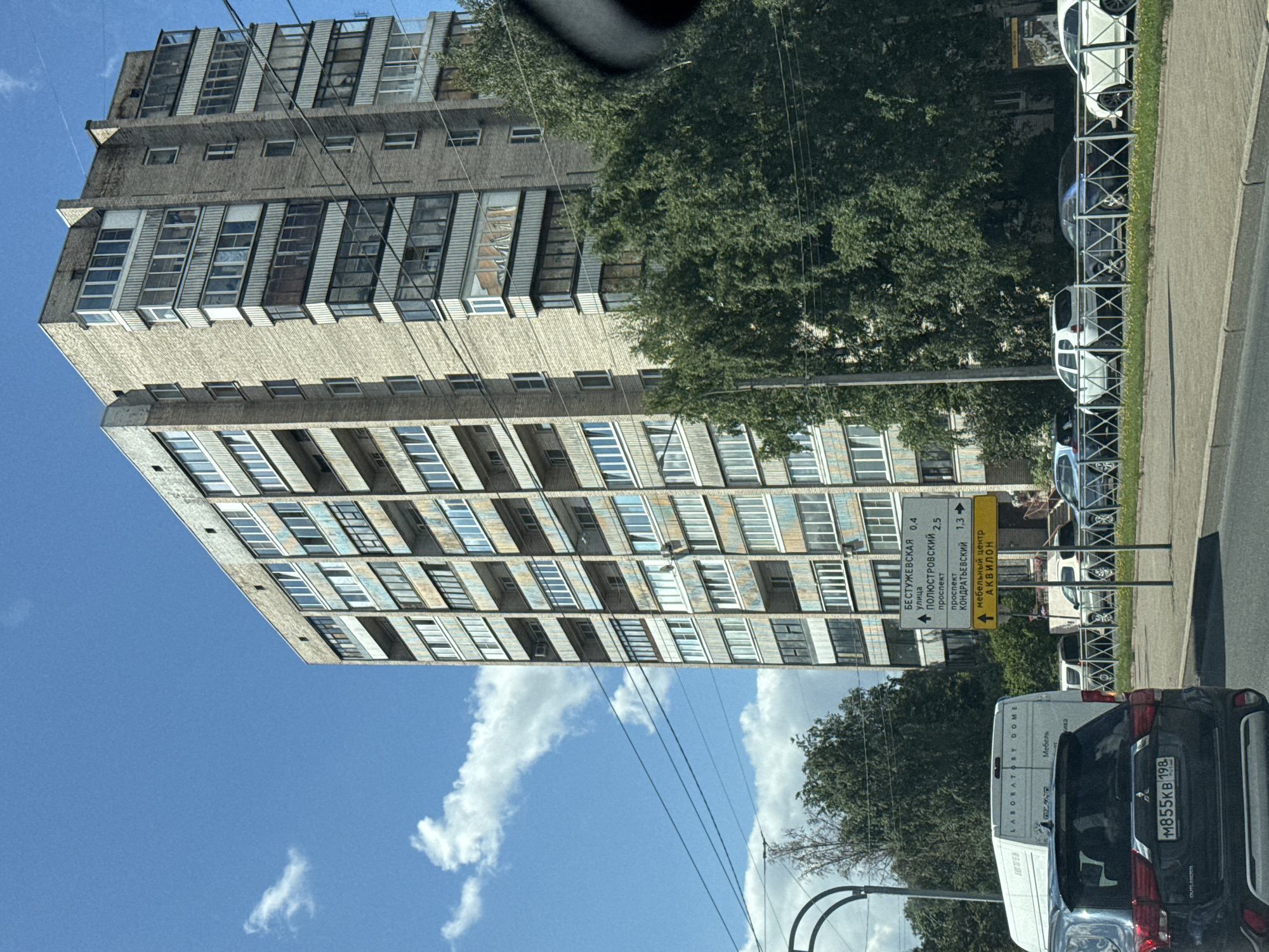 In Saint Petersburg, Russia, on a clear day with scattered clouds, a tall, multi-story brick apartment building dominates the scene. The building, characterized by numerous windows and balconies, stands adjacent to a street with parked cars and a white van. A street sign indicates directions to "Бестужевская улица" (Bestuzhevskaya Street) and "Полюстровский проспект" (Polyustrovsky Prospekt), with distances marked. Lush green trees line the street, partially obscuring the lower floors of the building. The foreground features the rear of a dark SUV.