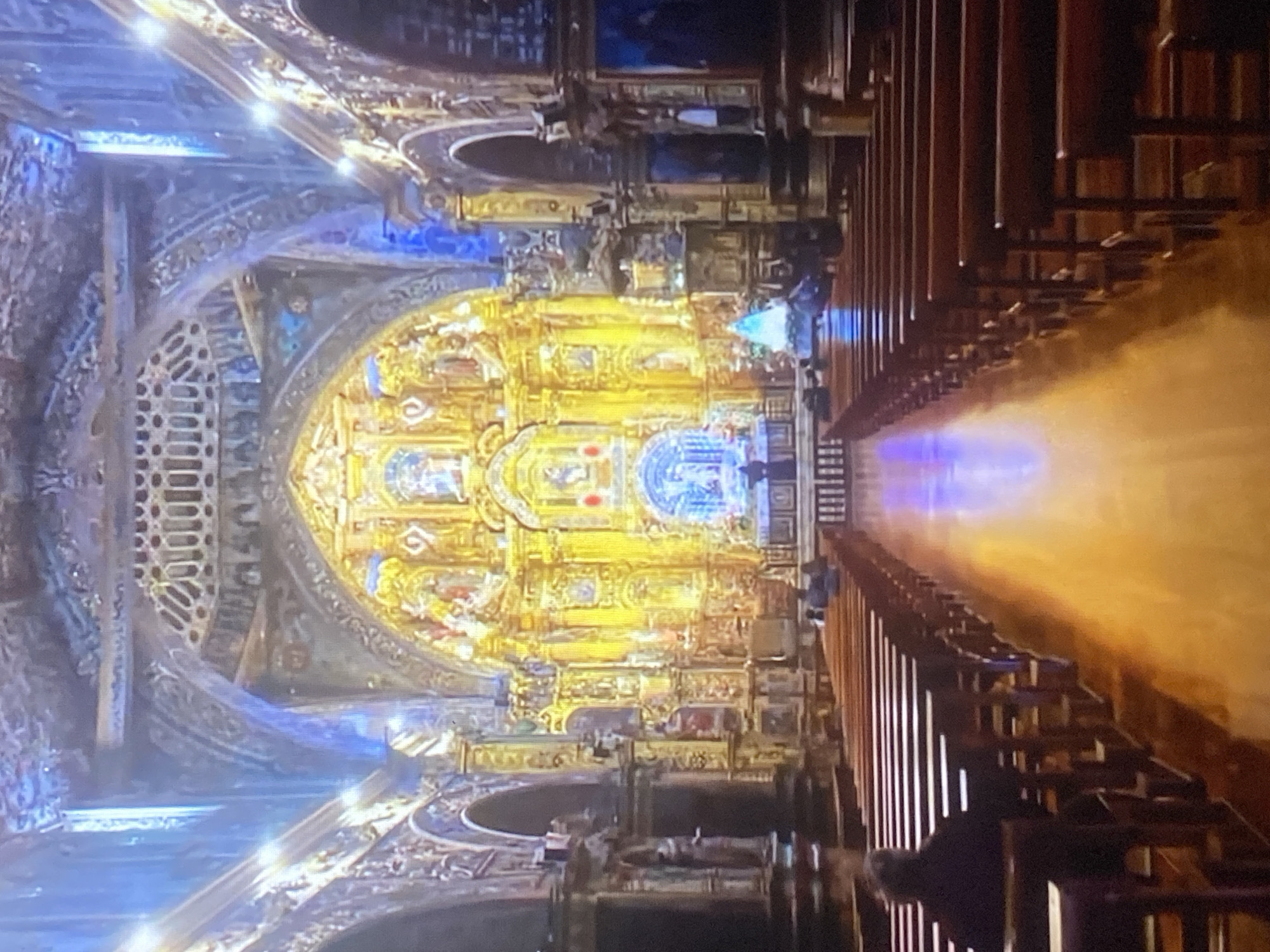 The image depicts the interior of the Iglesia de Tinaquillo, a church located in the city of Tinaquillo, Venezuela. The view is lengthwise down the nave, towards the altar and apse. The church is notable for its Baroque architecture and ornate decoration. The pews are visible on the right side of the nave and run towards the front. The altar is brightly lit and decorated with gold and blue motifs, which reflect the ornate decorations and sculptures of the apse. The church's vaulted ceiling is also elaborately decorated and illuminated by lights. Several people are standing or kneeling near the altar area.