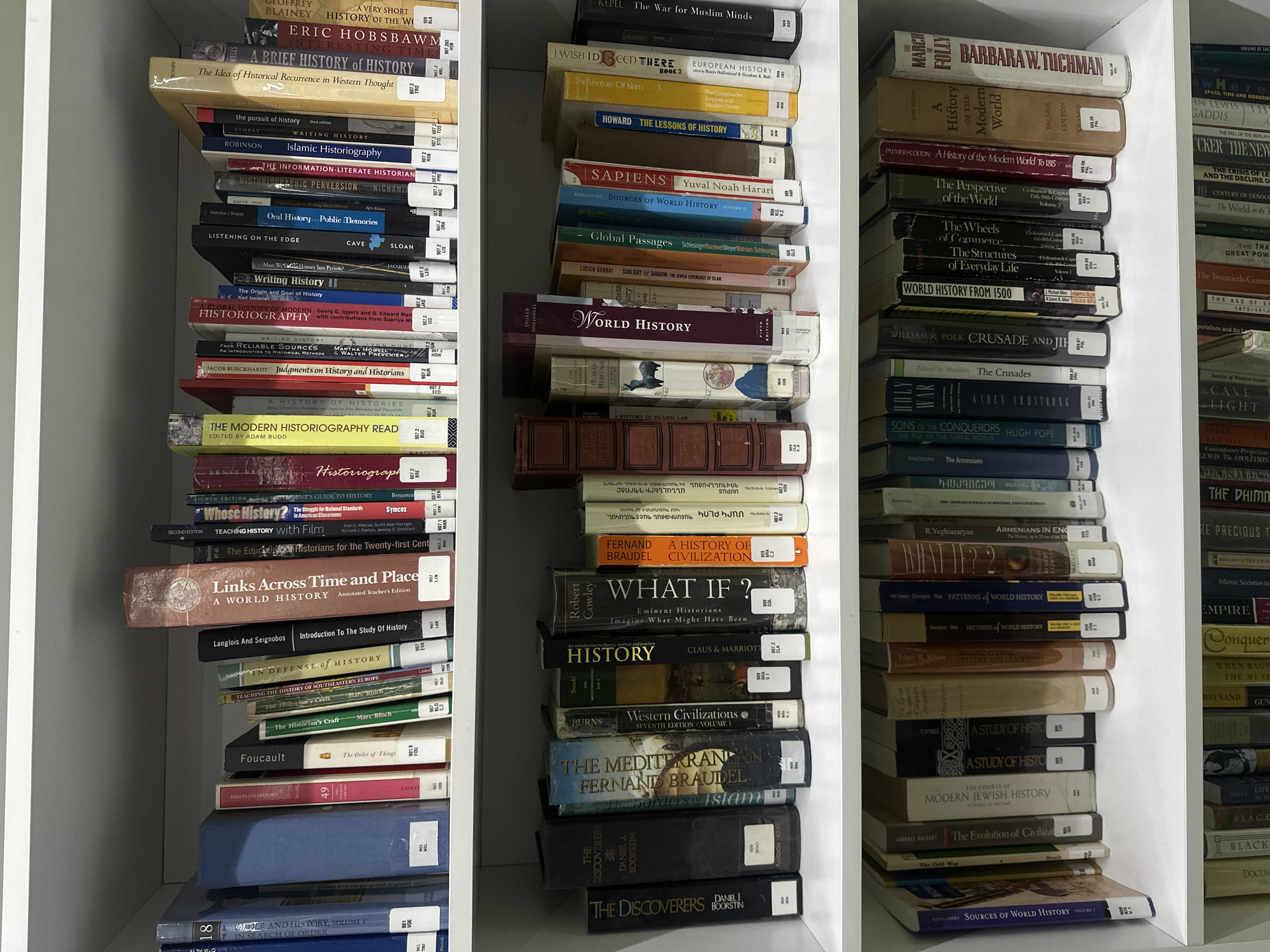 The image displays a white bookshelf filled with numerous books, primarily academic texts on history and historiography. The books are organized vertically in three main columns, with some shelves having books stacked horizontally or in small piles. Most books have white spine labels with black text, likely library call numbers, and some also have bar codes.
**Left Column (from top to bottom):**
The top shelf shows a book titled "A VERY SHORT HISTORY OF THE W" by GEOFFREY BLAINEY, followed by "ERIC HOBSBAWM INTERESTING TIME". Below this is "A BRIEF HISTORY of HISTORY" and "The Idea of Historical Recurrence in Western Thought".
The next shelf down includes books like "the pursuit of history" by STOREY, "Islamic Historiography" by ROBINSON, "THE INFORMATION-LITERATE HISTORIAN", "HISTORIOGRAPHIC PERVERSION" by NICHANTI, "Oral History Public Memories" by Hamilton | Shopes, "LISTENING ON THE EDGE" by CAVE, "Must We Do History Into Periods?" by JACQUES, "Writing History II", "The Origin and Goal of History" by Karl Jaspers, "GLOBAL HISTORY OF MODERN HISTORIOGRAPHY" by Georg G. Iggers and O. Edward Wan with contributions from Supriya Mu, and "WRITING HISTORY FROM RELIABLE SOURCES AN INTRODUCTION TO HISTORICAL METHODS" by MARTHA HOWELL. Also visible is "Judgments on History and Historians" by JACOB BURCKHARDT.
Further down, there's "A HISTORY OF HISTORIES" and "THE MODERN HISTORIOGRAPHY READ" edited by ADAM BUDD, followed by "Historiograph" by ERNST.
More books continue on lower shelves: "Whose History? The Struggle for National Standards in American Classrooms" by Symcox, "TEACHING HISTORY with Film" by Alan S. Marcus, Scott Alan Metzger, Richard I. Paxton, Jeremy D. Stod, "The Education of Historians for the Twenty-first Cent", "Links Across Time and Place A WORLD HISTORY", "Introduction To The Study Of History" by Langlois And Seignobos, "IN DEFENSE of HISTORY", "TEACHING THE HISTORY OF SOUTHEASTERN EUROPE", "The Historian's Craft" by Marc Bloch, "The Order of Things" by Foucault, and "ESSAYS ON HISTORY". The very bottom books include "DER AND HISTORY, VOLUME V" and "IN SEARCH OF ORDER".
**Middle Column (from top to bottom):**
The top book is "KEPEL The War for Muslim Minds". Below it, books include "I WISH IDEED THERE BOOK 2", "EUROPEAN HISTORY", "The Venture Of Islam 3", "The Gunpowder Empires and Modern Times", and "HOWARD THE LESSONS OF HISTORY".
Further down are "SAPIENS Yuval Noah Harari", "SOURCES OF WORLD HISTORY VOLUME I" by KEHLANISY, "Global Passages" by Schlesinger Blackwell Meyer Watrous-Schlesinger, and "SUNLIGHT & SHADOW, THE JEWISH EXPERIENCE OF ISLAM" by LUCIEN GUBBAY.
A prominent red-spined book is titled "WORLD HISTORY FIFTH EDITION", followed by other history texts including "A HISTORY OF ISLAMIC LAW", "THE HISTORY OF", and various volumes with Armenian titles or content, such as "ARMENIANS IN ENG The History, up to 20-ies of the XX" by R. Yeghiazaryan.
Lower down are "FERNAND BRAUDEL A HISTORY OF CIVILIZATION", "WHAT IF ? Eminent Historians Imagine What Might Have Been" by Robert Cowley, "HISTORY" by CLAUS & MARRIOTT, "Western Civilizations SEVENTH EDITION VOLUME 1" by BURNS, "THE MEDITERRANEAN FERNAND BRAUDEL", and "The borders of Islam".
The bottom books in this column are "THE DISCOVERERS" and "THE DISCOVERERS" by DANIEL J BOORSTIN.
**Right Column (from top to bottom):**
The books are partially visible due to being placed further back on the shelf. The top visible book is "THE MARCH OF FOLLY" by BARBARA W. TUCHMAN.
Below it, books include "A History of the Modern World" by PALMER COLTON (a multi-volume set, with "A History of the Modern World To 1815" visible), "The Perspective of the World Volume 3", "The Wheels of Commerce", "The Structures of Everyday Life Volume I", and "WORLD HISTORY FROM 1500".
Further down, titles like "CRUSADE AND JIH" by WILLIAM R. POLK, "The Crusades" edited by Madden, "HOLY WAR" by KAREN ARMSTRONG, "SONS OF THE CONQUERORS" by HUGH POPE, and "The Armenians" by PANOSSIAN are visible.
Below these, books include "PATTERNS of WORLD HISTORY" by von Sivers Desnoyers Stow (multiple volumes), "ORGANIZATIONAL CULTURE AND LEADERSHIP" by Edgar H. Schein, "A STUDY OF HISTO" by TOYNBEE (appearing twice), "THE COURSE OF MODERN JEWISH HISTORY" by HOWARD M. SACHAR, "The Evolution of Civilizat" by CARROEL BRILLER, "The Cold War", and "SOURCES OF WORLD HISTORY VOLUME I" by KOHLANSKY.
The very bottom of the right column shows "SOURCES OF WORLD HISTORY VOLUME I".
The books appear to be from a personal library or a specialized academic collection, given the consistent subject matter of history, historiography, and world civilizations. Many books have colored spines (red, blue, green, brown) and show varying degrees of wear.