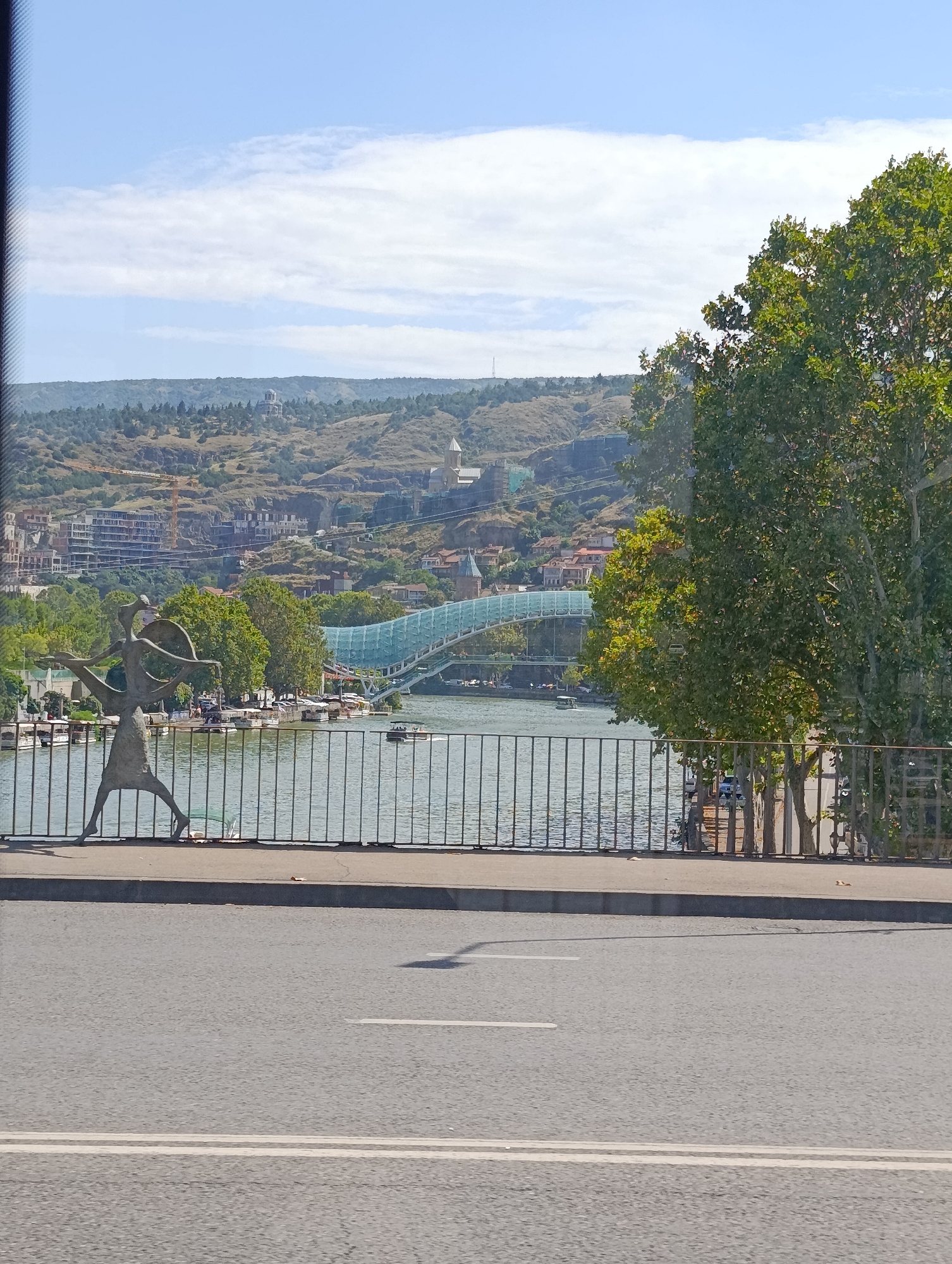 The image depicts a panoramic view of the Mtkvari River in Tbilisi, Georgia, with the Bridge of Peace prominently featured. In the foreground, a pedestrian walkway is visible with a metal railing, and a bronze sculpture of a human figure in a dynamic pose. Beyond the railing, the Mtkvari River flows, with several boats docked along its banks. The Bridge of Peace, a modern pedestrian bridge made of glass and steel, spans the river. In the background, the hilly landscape of Tbilisi rises, dotted with buildings and trees. A prominent church or cathedral with a dome is visible on one of the hills. The sky is blue with scattered white clouds, and the overall lighting suggests a bright, sunny day. The scene appears to be a public space, likely a popular tourist spot.