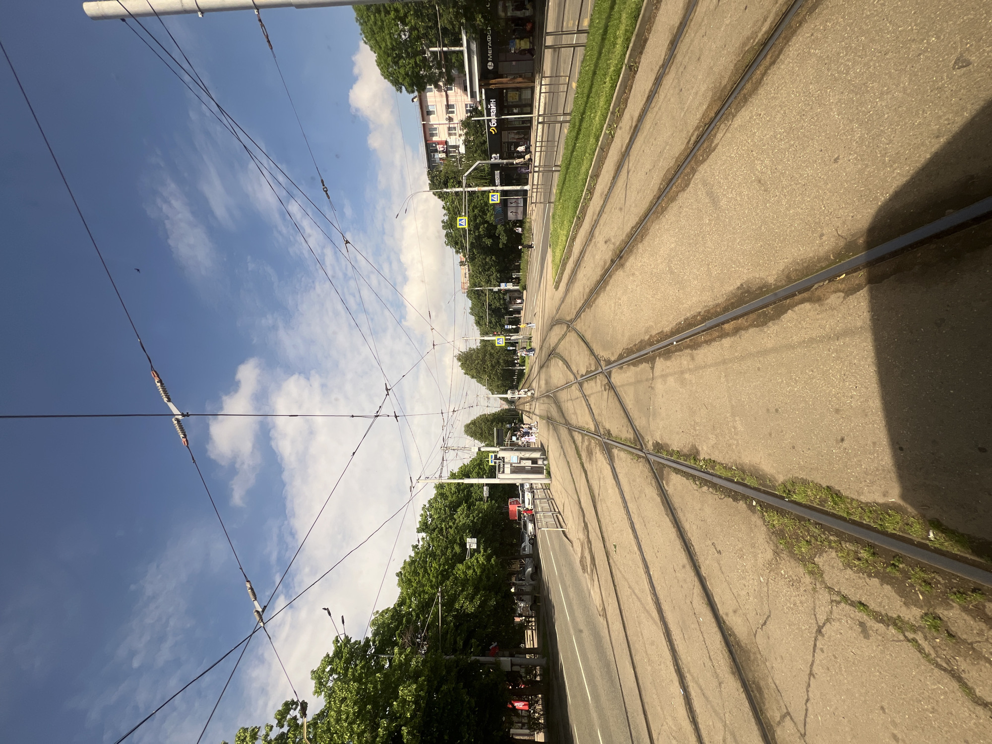 The image shows a street in Krasnodar, Russia, with tram tracks running along the road. The perspective is from a low angle, possibly from within a tram or near the tracks. The sky is visible with scattered clouds, and overhead power lines are prominent. Buildings and trees line the street, and pedestrian crossings can be seen in the distance. Some individuals and vehicles are present but not clearly identifiable. The road and tracks show signs of wear and tear.