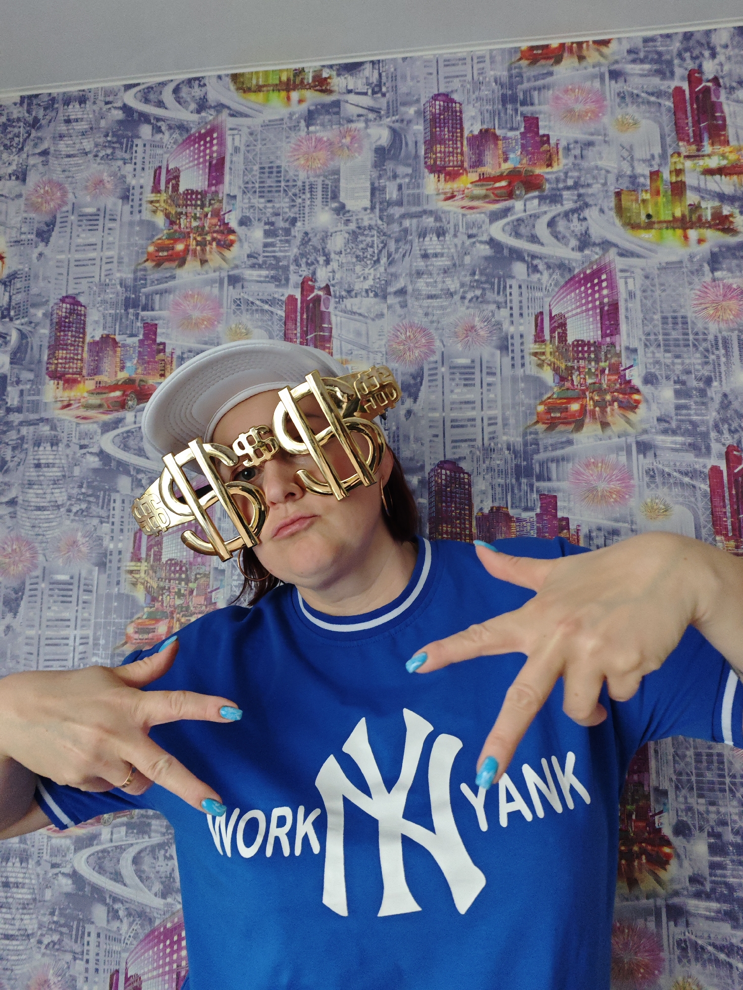A person wearing a blue t-shirt with "WORK YANK" printed on it, along with oversized gold dollar sign glasses and a white cap, is posing against a wallpapered backdrop featuring images of a city with highways and buildings. The person is making hand gestures with painted blue fingernails. The location is Марковский, Russia.