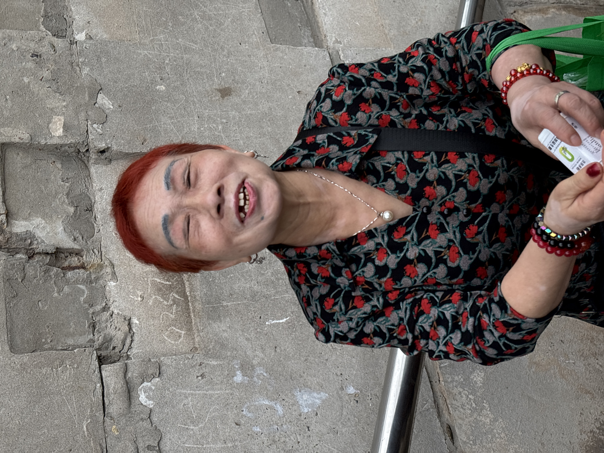 The image shows an older woman with red hair standing on gray concrete steps in an outdoor setting. She is wearing a black floral print top with red and gray flowers, a black strap across her chest, and multiple beaded bracelets on her right wrist. She is holding a small white object in her hands. She has a silver necklace with a pearl and blue eyebrows. She appears to be smiling, and her teeth are visible. The location appears to be in Jiangjin, China.