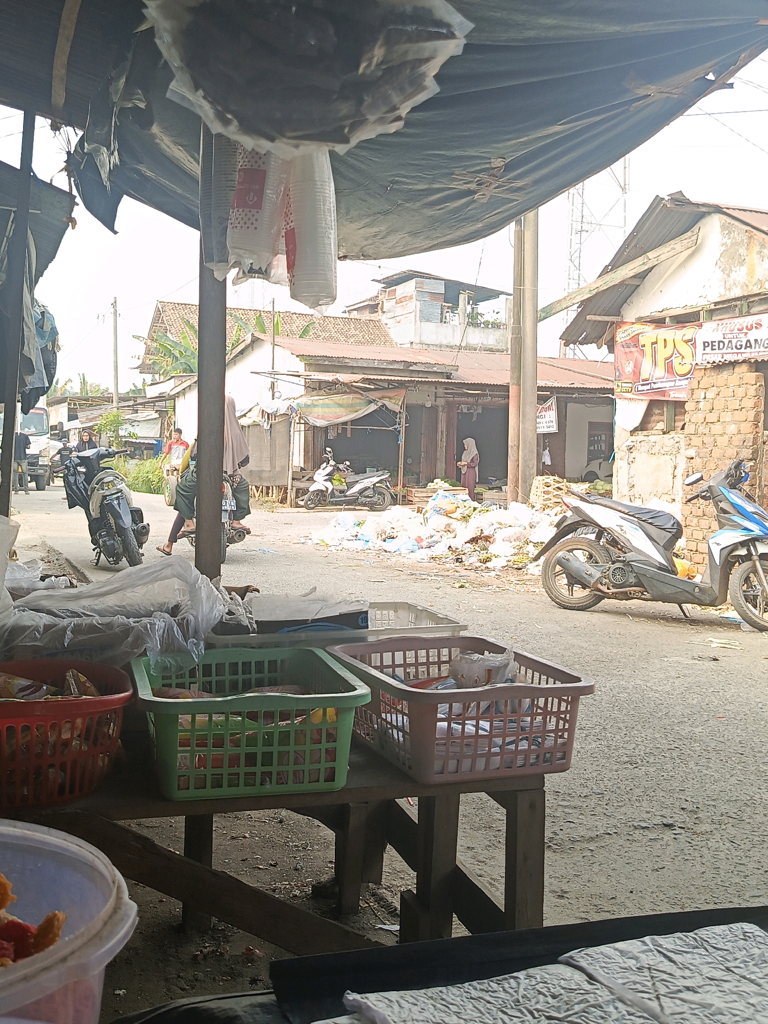 This is a ground-level, eye-level photograph taken outdoors in what appears to be a marketplace in Megang Sakti I, Indonesia. The foreground is dominated by plastic storage baskets, one red and one green, filled with various items, and a clear plastic container with some red and orange objects. A wooden table supports the baskets. Beyond the table, the scene opens to a street with several motorcycles parked. In the middle ground, a pile of trash or debris is visible on the ground. Several buildings line the street, some with brick construction and corrugated metal roofs. A few people are present; one person wearing a light-colored hijab stands near a building on the left, and another person in a similar head covering is on the back of a motorcycle in the center of the image. Another person is riding a bicycle further down the street. The sky is bright, suggesting a daytime scene. Tarps are visible overhead, providing shade over some stalls. Signs on the buildings are in Indonesian.