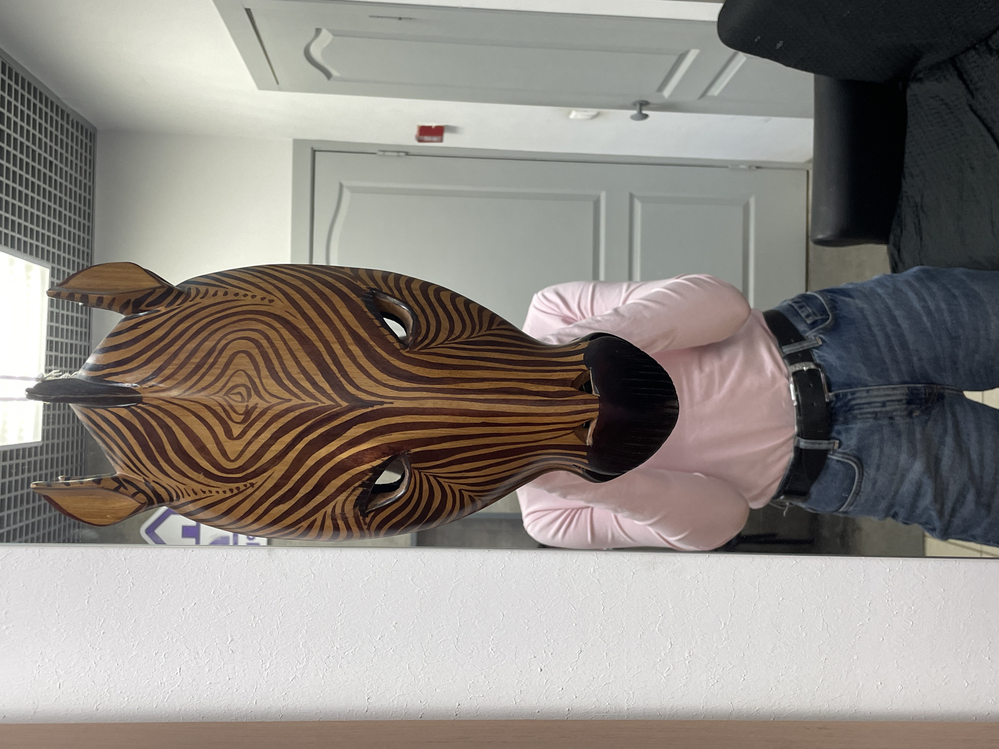 An indoor scene in Izhevsk, Russia, depicts a single individual holding a large wooden zebra mask directly in front of their face, obscuring it entirely. The person wears a light pink long-sleeved shirt, blue denim jeans, and a black belt with a silver buckle, with their hands gripping the lower sides of the mask. The mask is intricately carved from wood, featuring light and dark brown stripes mimicking a zebra's pattern, prominent ears, and carved eye openings.
The background shows a brightly lit interior space with light-colored, possibly textured, walls. A suspended grid ceiling with recessed lighting is visible overhead. Two light gray interior doors with paneling are present in the background, one fully visible to the right of the mask and another partially visible behind the mask. A red rectangular object is affixed to the wall between the two doors. A portion of a black upholstered armchair or sofa is visible in the lower right corner of the frame. The overall environment suggests an office, waiting area, or commercial hallway.