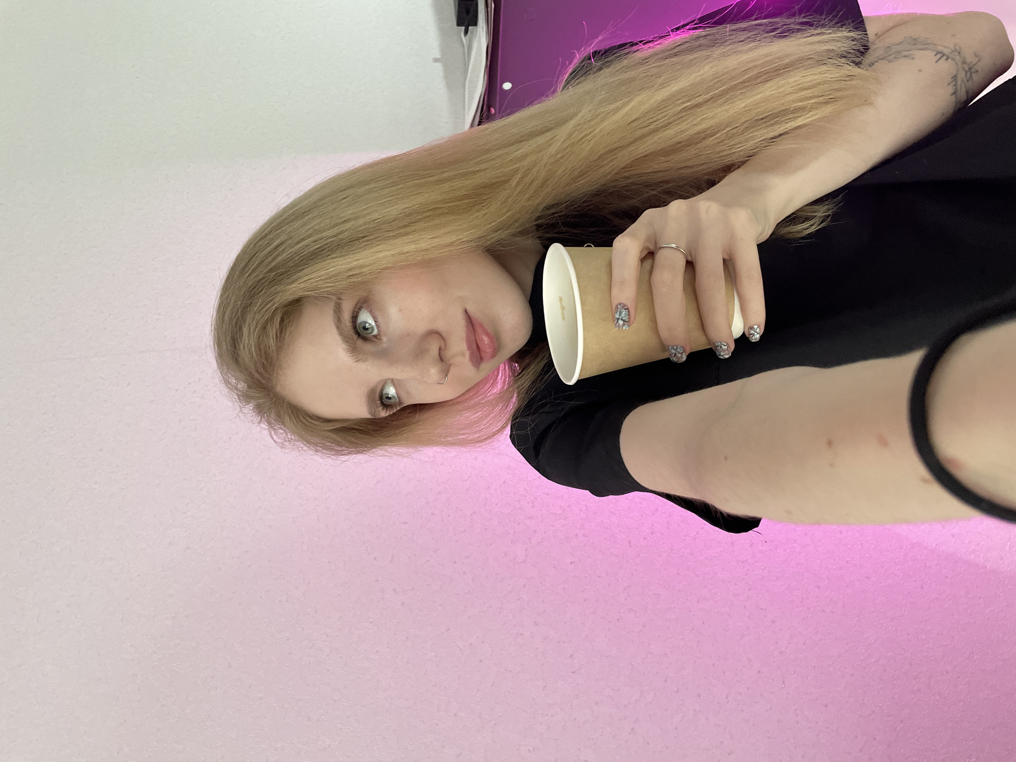 A fair-skinned young woman with long blonde hair is holding a disposable paper coffee cup with a black lid. She has pale skin and blue eyes. She is wearing a black top. She has a nose ring. She has a tattoo on her forearm. She is wearing a ring on her finger. Her fingernails are decorated with a floral design. The background is a light pink wall. There is an object on the wall above her head. The picture appears to be taken in an indoor setting. There is no verifiable information about Izhevsk, Russia in the image.