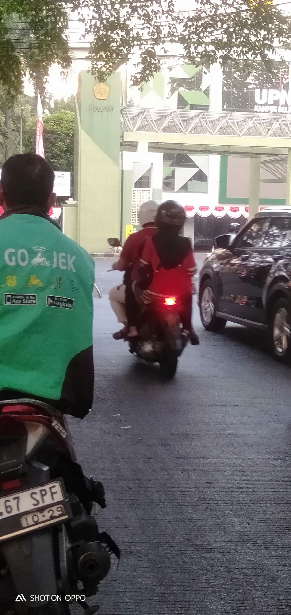 In South Jakarta, Indonesia, a man wearing a green Gojek jacket is facing away from the camera, suggesting he is either an observer or a Gojek driver. He stands on a paved road, likely a street, with a motorcycle visible in the foreground and a black SUV in the background. To the left, a large green pillar with the text "UPNVJ" is visible, indicating the location might be near the Universitas Pembangunan Nasional Veteran Jakarta campus. Behind the pillar, a flag on a flagpole can be seen. The overall scene depicts a street environment with urban infrastructure, suggesting a typical day in South Jakarta.