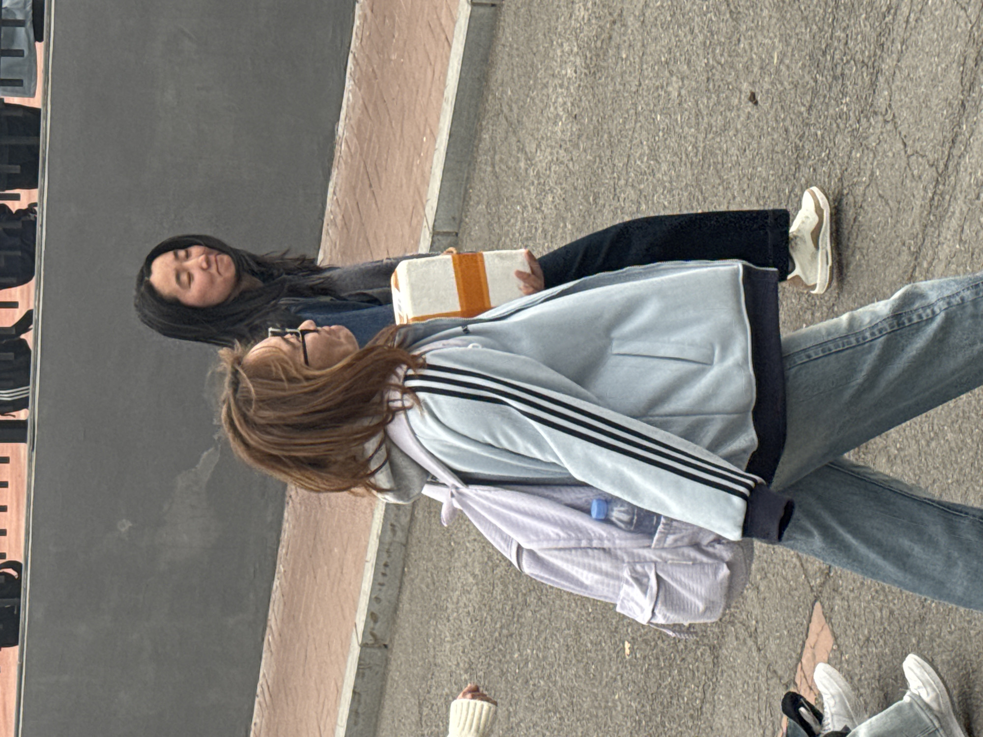 The image depicts two young women outdoors in Zhongtun, China. One woman with long, dark hair is lying down, holding a white package with orange tape. She is wearing dark clothing and light-colored sneakers. The other woman, standing next to her, has brown hair and is wearing glasses, a light blue Adidas jacket, blue jeans, and a light purple backpack. The scene takes place on a paved area, possibly a sidewalk, adjacent to a grey wall and a brick-paved area.