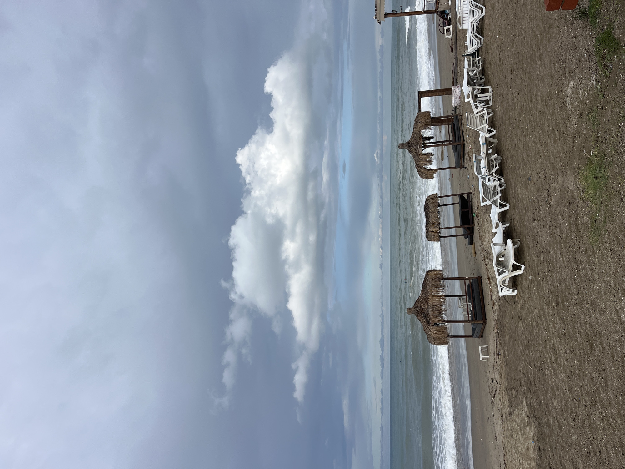 The image shows a beach in Alanya, Turkey, on a cloudy day. The beach is lined with straw-roofed cabanas and white plastic chairs. The sea is visible, with waves breaking on the shore. The sky is overcast, with large cumulus clouds dominating the view. No people are visible in the shot.