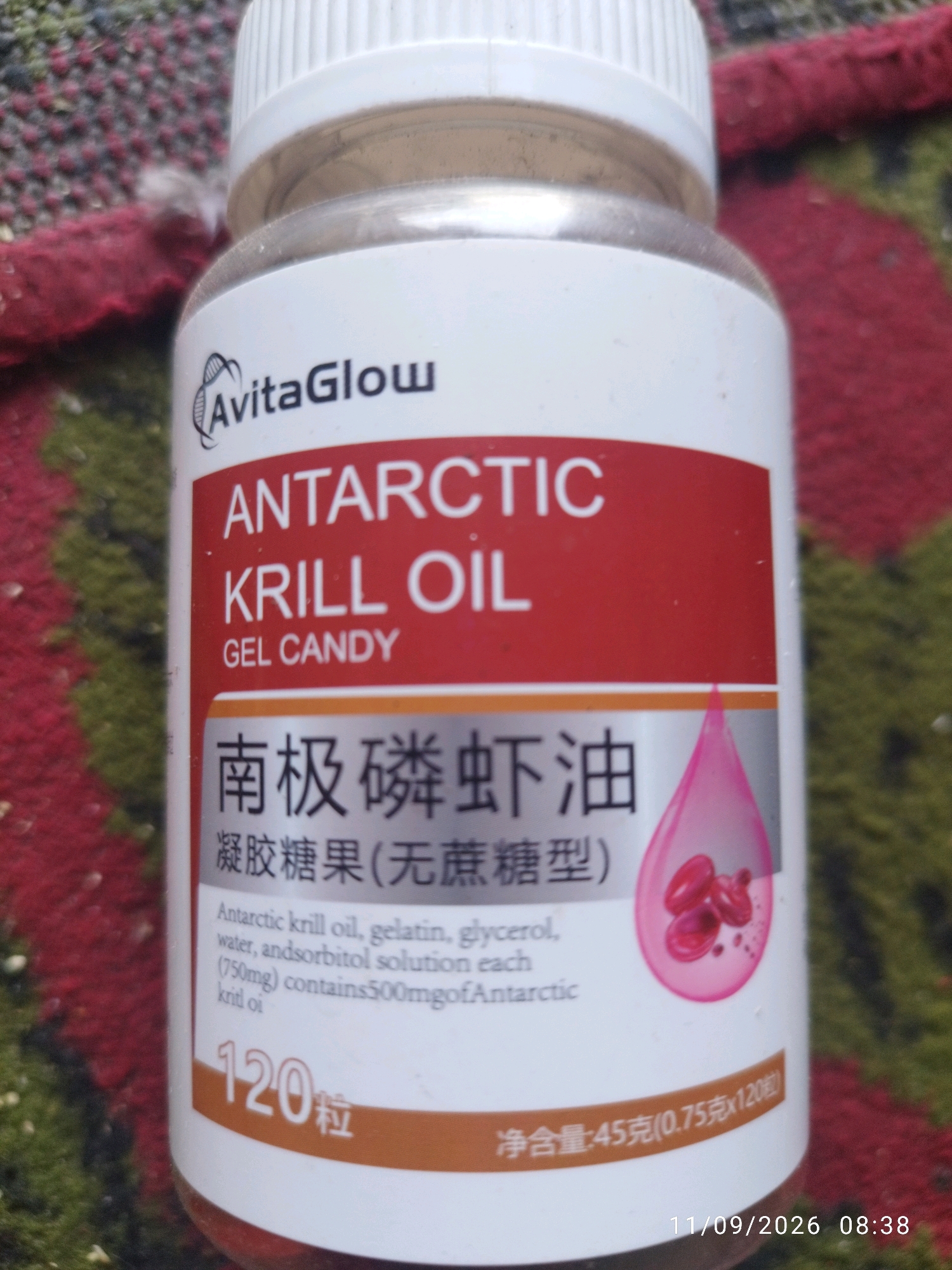 The image shows a bottle of AvitaGlow Antarctic Krill Oil Gel Candy. The label is primarily white with red and orange accents. The brand name "AvitaGlow" is prominently displayed at the top. Below it, "ANTARCTIC KRILL OIL" is written in large white letters on a red background, followed by "GEL CANDY". There is Chinese text below this, which translates to "Antarctic Krill Oil". A pink droplet with three red capsules inside is depicted on the right side of the bottle. The ingredients list, in English, states "Antarctic krill oil, gelatin, glycerol, water, andsorbitol solution each (750mg) contains 500mg of Antarctic krill oi". At the bottom left, "120 粒" is written, indicating 120 capsules. The bottom right shows "净含量:45克(0.75克x120粒)" which translates to "Net content: 45g (0.75g x 120 capsules)". The bottle is placed on a textured surface that appears to be a blanket or rug with red and dark green colors. The lighting suggests it is indoors or in a shaded outdoor area. A timestamp "11/09/2026 08:38" is visible at the bottom right of the image, likely from a camera or mobile device. There are no people, animals, or any elements that would place this image in Mojokerto, Indonesia.