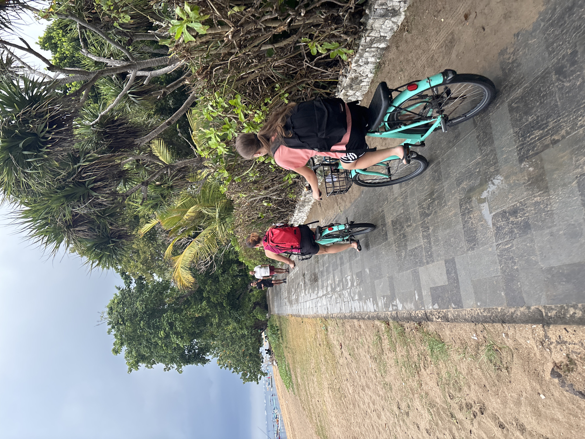A group of individuals are on a paved pathway adjacent to a sandy beach. Two young females are riding turquoise bicycles. The female in the foreground has a black backpack and is wearing a coral-colored shirt and black shorts. The female behind her has a red backpack and is wearing a black tank top and shorts. Two adults, a male and a female, are walking on the pathway ahead of the cyclists. The beach to the left has scattered green vegetation and numerous small boats anchored in the ocean. The pathway is lined with lush green trees and dense bushes. The sky is overcast.