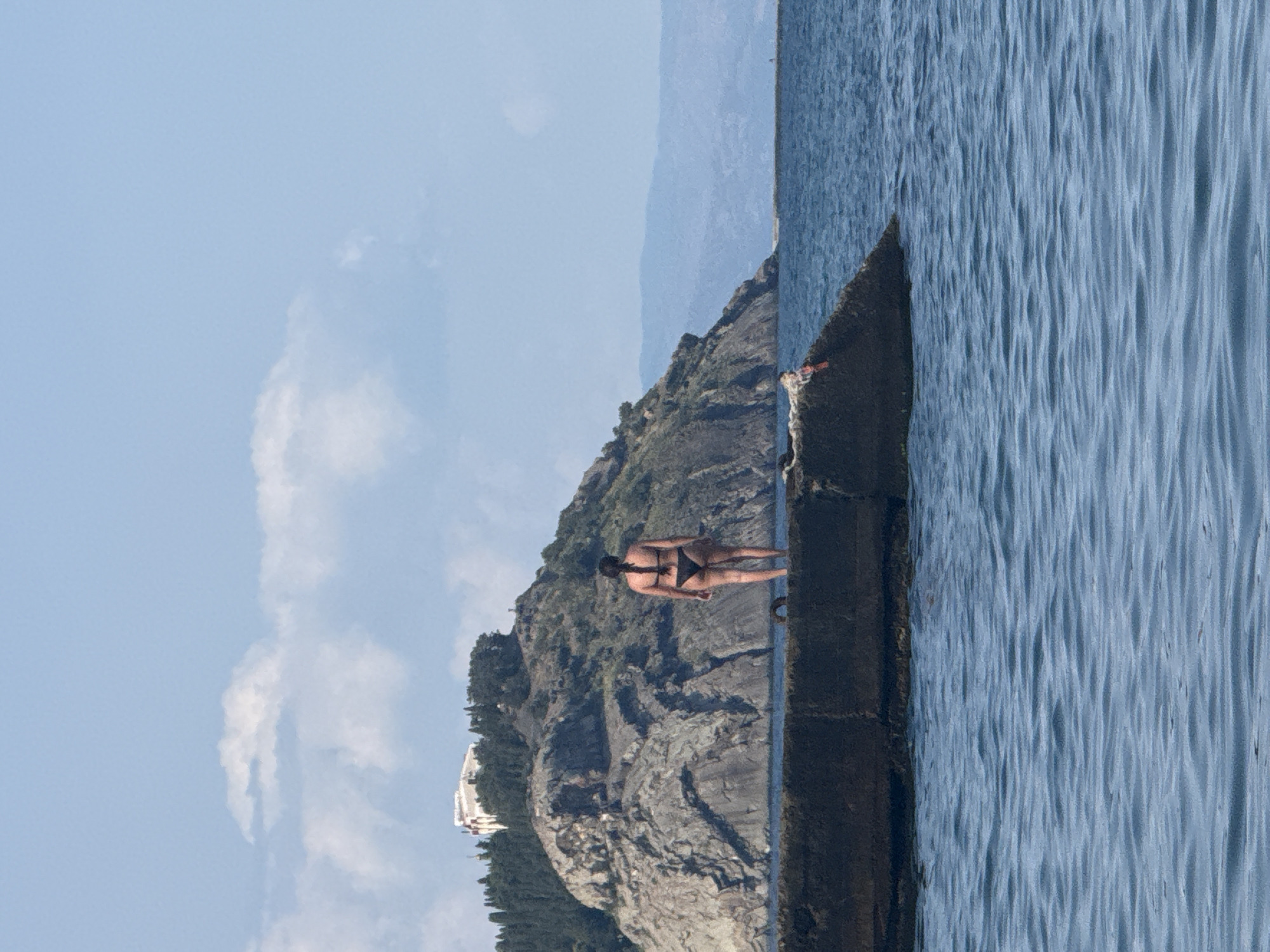A woman in a black bikini stands on a concrete pier facing away from the camera, looking out at the sea. Behind her, a rugged, tree-covered mountain rises from the water. A white building is visible on the mountaintop. The sea is calm with light blue waves. The sky is a clear light blue with a few scattered white clouds. The scene appears to be set in a coastal area, possibly Partenit, Ukraine.