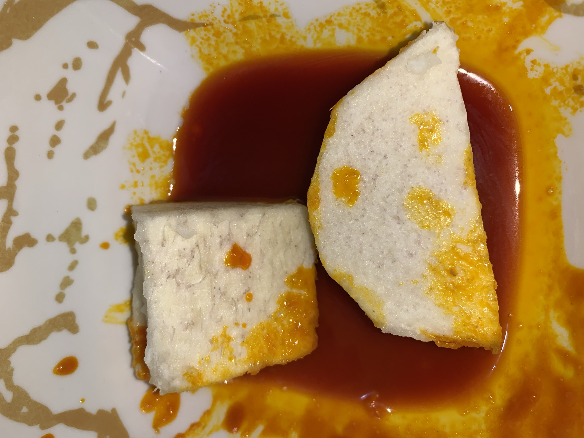 Two pieces of what appears to be food, possibly taro or yam, are on a white plate with an abstract gold pattern. The pieces are covered in a red and yellow sauce. The scene is indoors, and judging from the plate and the food, it is likely a dining setting. The location in Rafin Par, Nigeria is not evident from the image itself, so its inclusion must be inferred or known from external information.