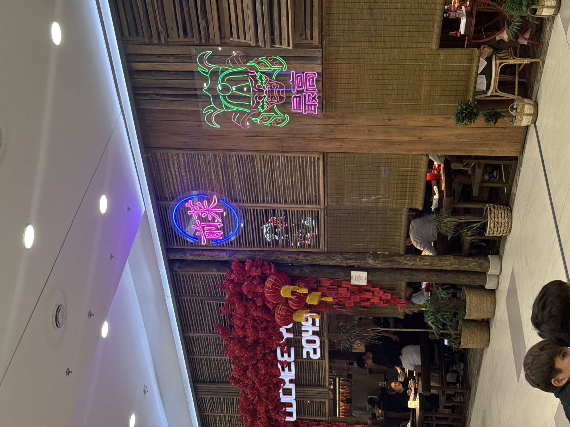 The image captures the interior of a restaurant with a distinct East Asian theme, possibly located in Almaty, Kazakhstan, given the context. The restaurant features a rustic wooden interior with slatted wood panels and bamboo blinds. A prominent neon sign in bright pink reads "LUCHEEE Y" followed by "2049", with a large arrangement of artificial red flowers and red lanterns above and to the left. To the right, another neon sign glows in green and pink, depicting a stylized dragon-like face and Chinese characters below it. Several tables with diners are visible in the background, some seated on benches. Potted plants in woven baskets are placed strategically around the dining area. The floor is tiled. Two young individuals are partially visible in the foreground, their faces turned away from the camera, suggesting they are seated at a table. The lighting in the scene is a mix of ambient restaurant lighting and the bright glow of the neon signs, with some purple and white accent lighting from the ceiling.