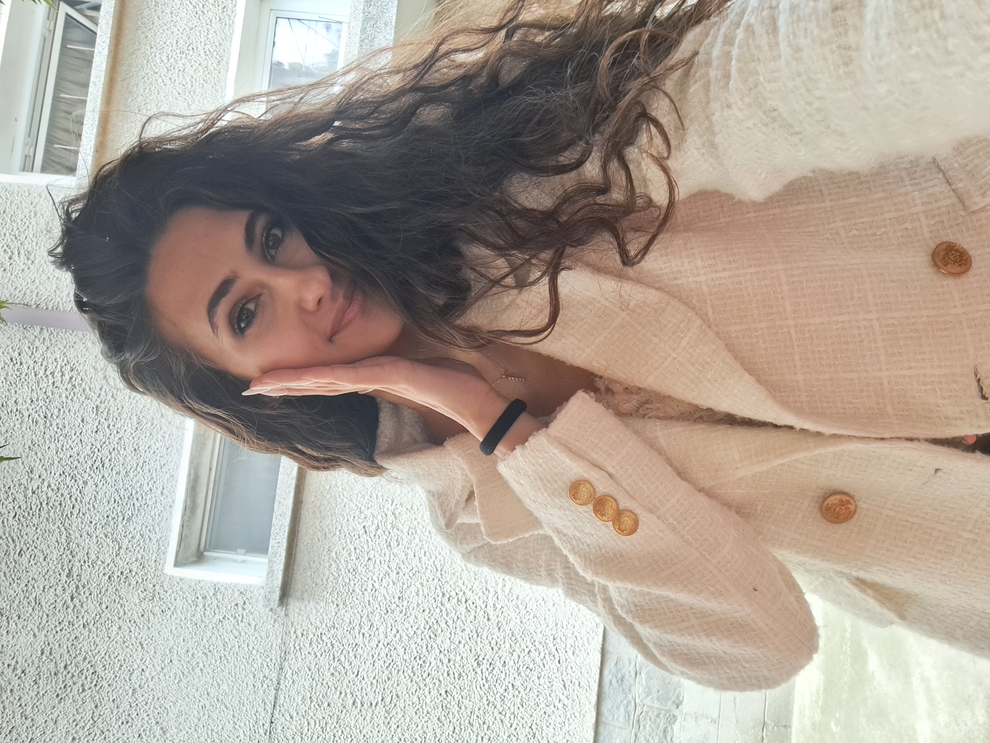 A young woman with long, dark, curly hair and dark eyes is posing for a selfie outdoors. She is wearing a cream-colored tweed blazer with gold buttons. Her right hand is resting on her cheek, and she is looking directly at the camera with a slight smile. Behind her, there is a textured off-white building with two windows. The scene appears to be in a residential area, potentially Negovan, Bulgaria.