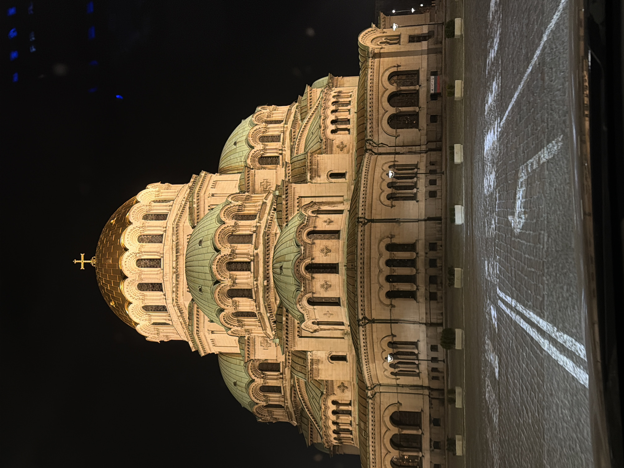 An exterior long shot captures the Alexander Nevsky Cathedral in Sofia, Bulgaria, at night, illuminated against a dark, cloudless sky. The massive Neo-Byzantine structure dominates the frame, featuring a central golden dome topped with a prominent golden Orthodox cross. Surrounding the central dome are numerous smaller domes, all featuring a distinct pale green or turquoise color, tiered and connected to the light-colored stone facade. The cathedral's exterior is characterized by multiple arched windows and decorative architectural elements.
In the foreground, a cobblestone plaza or road surface is visible, reflecting the ambient light and displaying clear white double parallel lines. A fainter white arrow, pointing left, is also discernible on the pavement. The bottom edge of the image reveals the top of a vehicle's dashboard or windshield, indicating the photograph was taken from inside a car. Along the base of the cathedral grounds, four evenly spaced white rectangular planters containing dark green shrubs are visible. Several streetlights emit light in the mid-ground. No individuals are present in the scene. A small, blurry rectangular object with red, blue, and white stripes, possibly a flag, is partially visible near the base of the cathedral wall behind a planter on the right side.