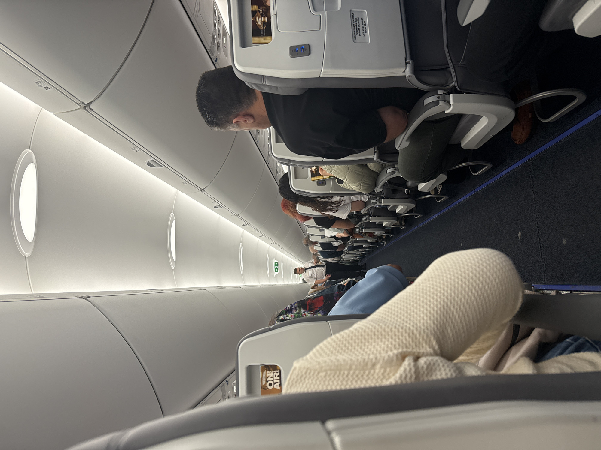 The image shows the interior of an airplane cabin. Passengers are seated in rows along the aisle. A man in a black shirt is seated in the foreground. There's a flight attendant visible down the aisle. The seats have headrests and personal screens. There are overhead storage compartments above the seats. A sign on the seat in front reads "ON AIR", followed by Cyrillic text and English translation stating "LIFE VEST UNDER YOUR SEAT." The plane's interior is primarily white and grey. The city of Sofia, Bulgaria cannot be determined from this image.