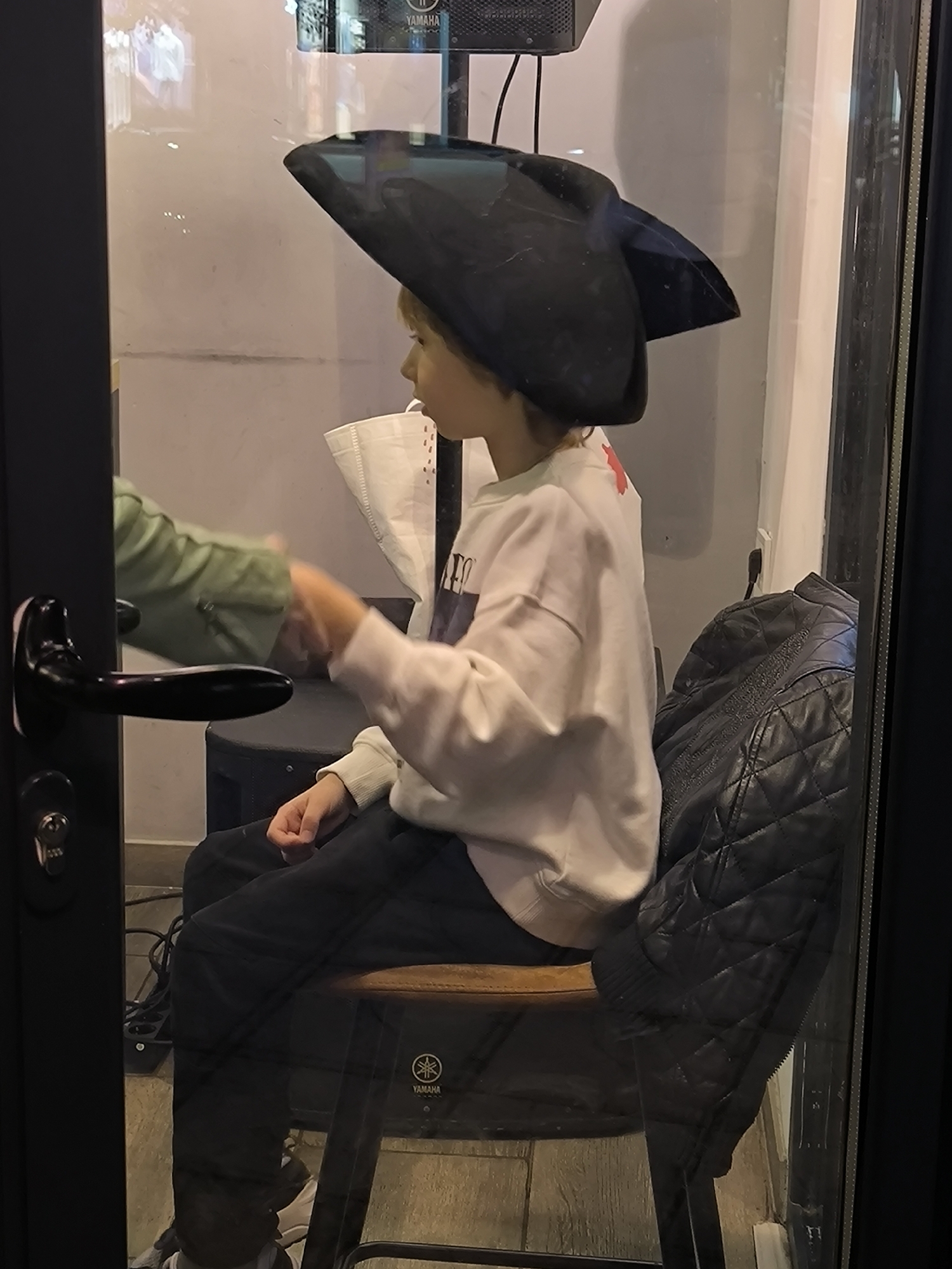 A young individual is seated in profile on a wooden stool within a glass-enclosed space, identified as being in the city of Bar, Montenegro. The individual, appearing to be a child or pre-teen, wears a dark tricorn-style hat, a light-colored long-sleeved sweatshirt, and dark trousers, with white sneakers visible. Their right hand is extended, making contact with the hand of a second, partially visible individual, whose arm is clad in a green long-sleeved garment, entering the frame from the left through an open black door frame. The door frame features a black handle and a visible key in a lock cylinder.
Behind the seated individual, a black quilted jacket is draped over the back of the stool or an adjacent chair. A white, crinkled bag or piece of fabric is positioned behind their head. Above and to the right, a black speaker unit bearing a "YAMAHA" logo is mounted on the light-colored wall, with connected wires. Another "YAMAHA" logo is visible on the metal leg of the wooden stool. The floor consists of light brown material, possibly tiles or laminate. Reflections are present on the glass enclosure, suggesting external lighting or elements. The interaction between the two hands appears to be a greeting or an exchange.