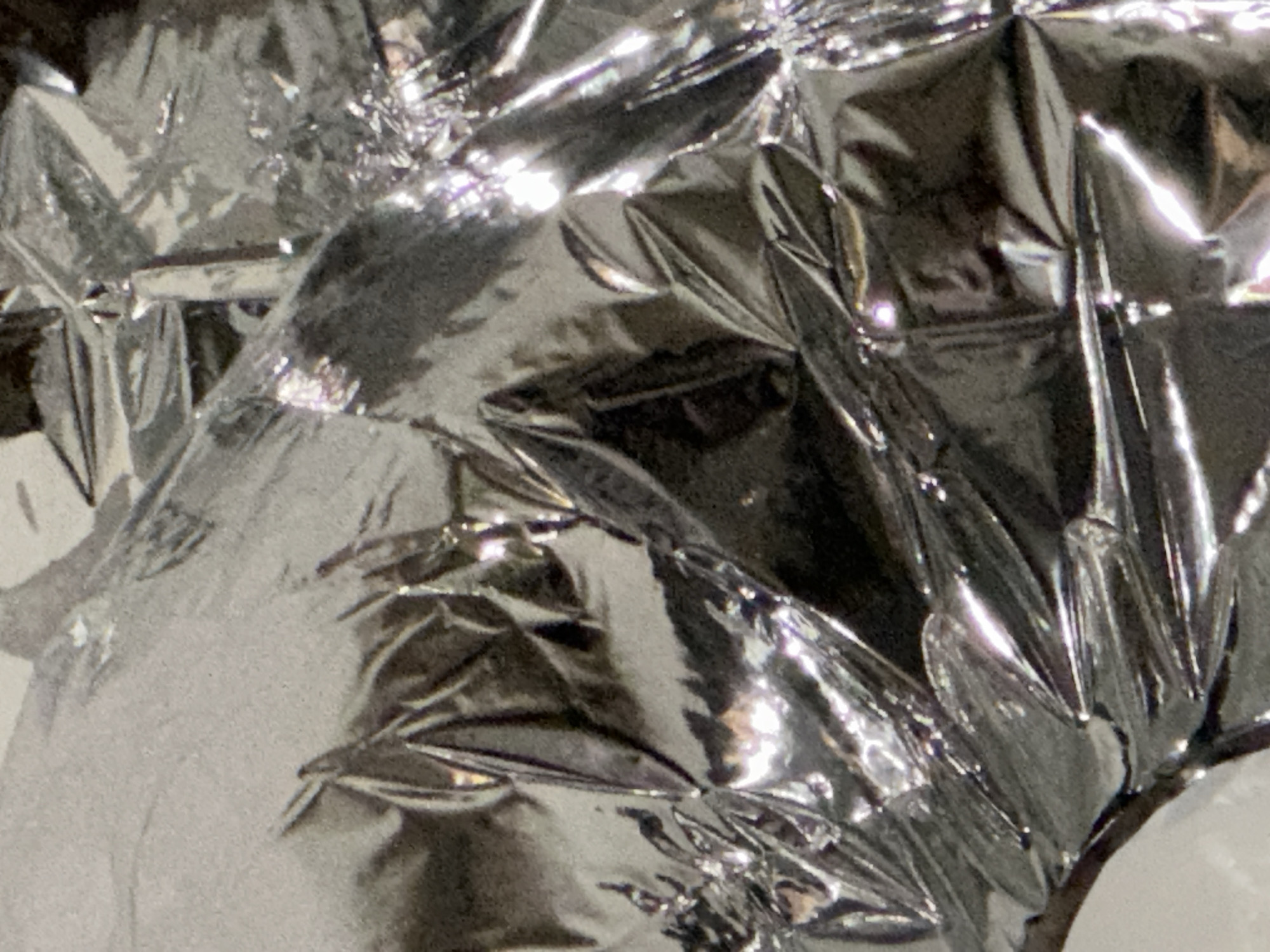 The image presents a close-up, abstract view of a heavily crumpled, highly reflective silver-colored material, consistent with metallic foil or metallized plastic sheeting. The surface is characterized by numerous sharp creases, folds, and irregular wrinkles that create a complex, three-dimensional texture. Bright, elongated specular highlights are present across the material, indicating reflections from an unseen light source, contrasting with darker, shadowed areas that define the contours and depths of the crumpled surface. No people, discernible objects, or specific environmental features are visible within the frame, and no actions or interactions are depicted. This scene is located in the city of Tunbridge Wells, United Kingdom.