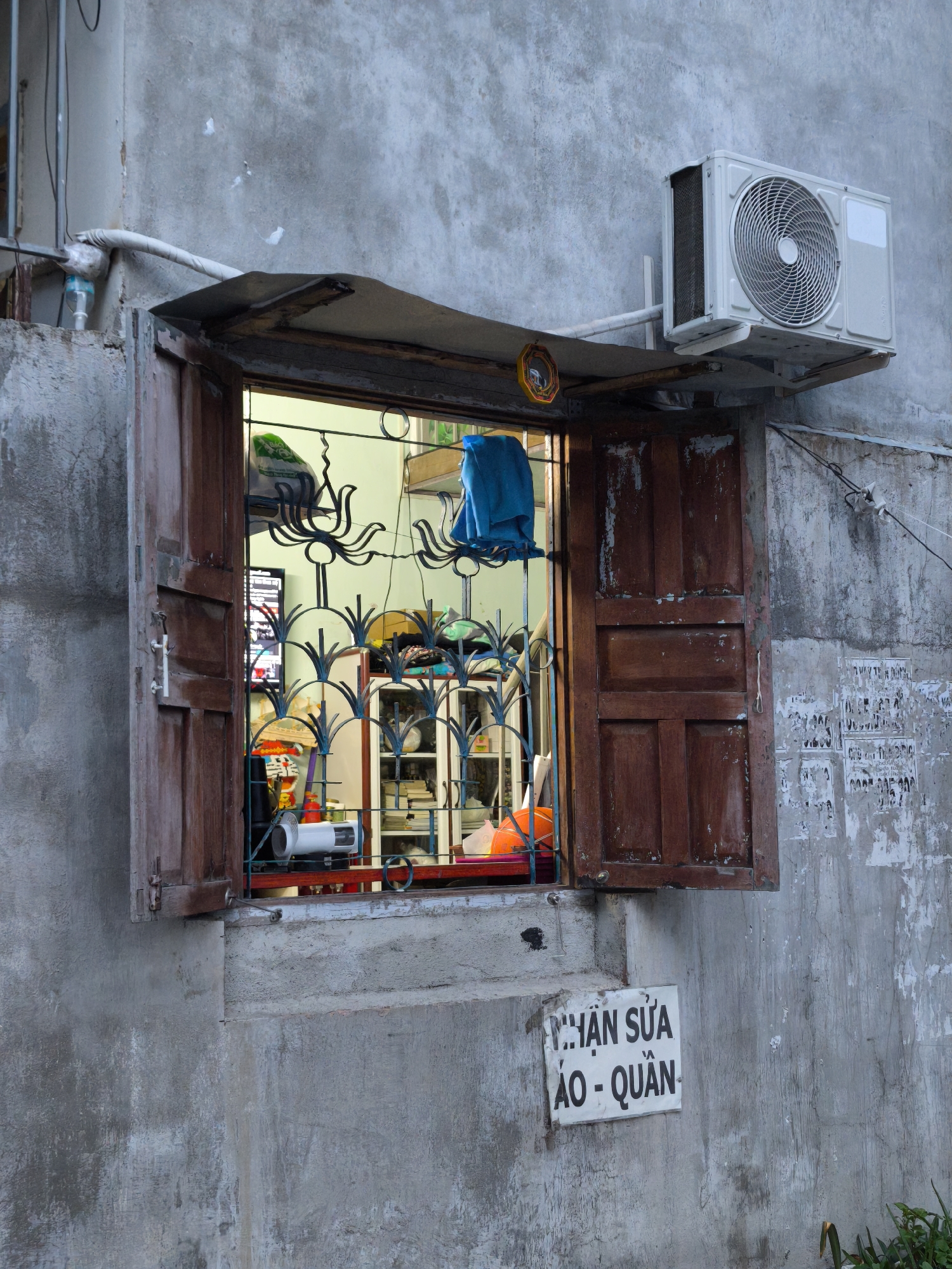 The image shows the exterior of a weathered concrete building, likely an apartment or shop, in Bac Nha Trang Ward, Vietnam. An open wooden-shuttered window reveals a cluttered interior with a metal security grate. Inside, various items are visible, including what appears to be a television, shelves stocked with books and objects, a blue garment hanging, and a white electric fan. An air conditioning unit is mounted on the exterior wall above the window. Below the window, a sign with Vietnamese text is affixed to the concrete wall. The overall impression is one of a utilitarian and lived-in urban environment.