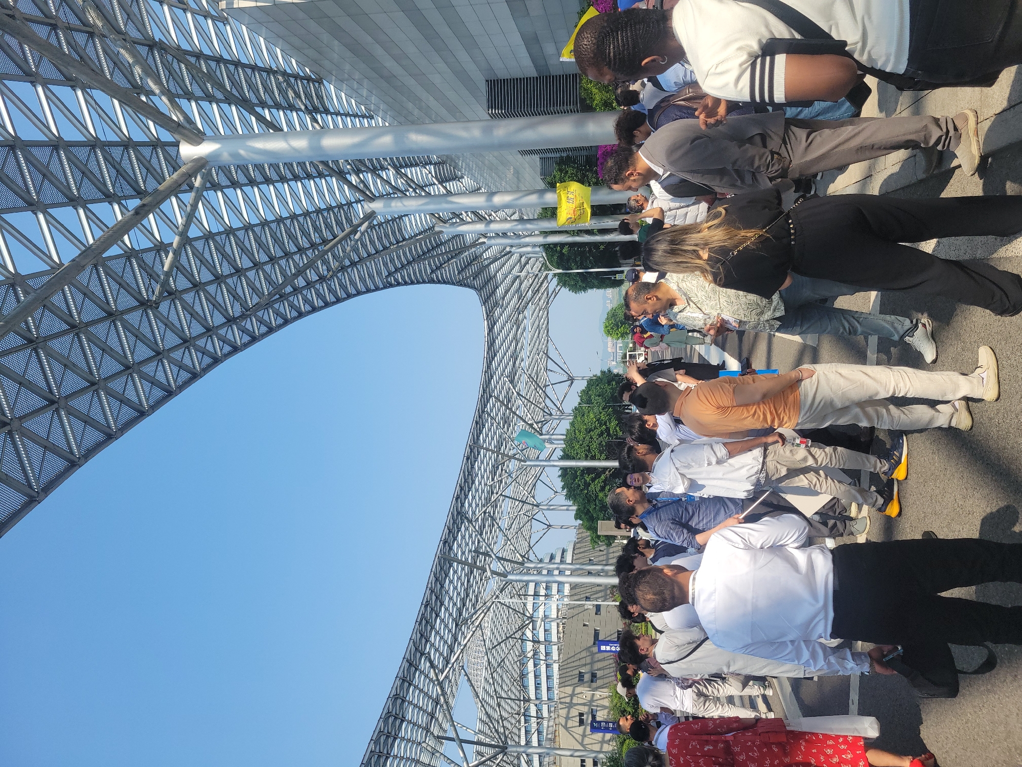 The image shows a crowd of people standing under a large, arched structure made of metal beams, with a clear blue sky visible above. The people are of various ethnicities and are wearing a range of casual and business attire. They appear to be in a line or gathering, possibly waiting for something. The location appears to be an urban environment, with buildings and trees visible in the background. The presence of signs written in Chinese suggests the city of Liangjiang, China, as the possible location. The modern architecture of the structure and the surrounding environment suggests a contemporary setting.
