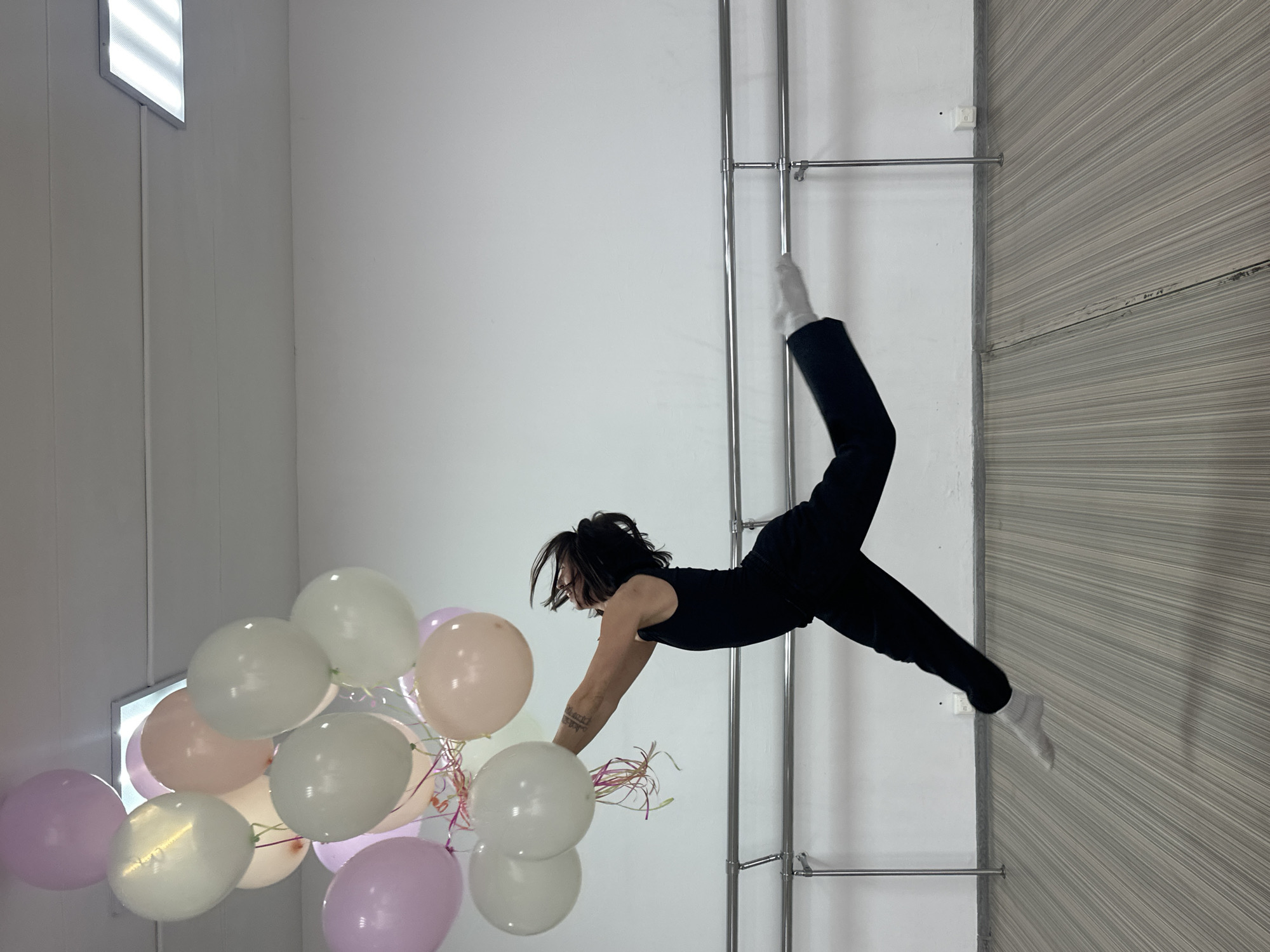 The image depicts a person in a black sleeveless top and black pants suspended horizontally in a room with white walls. The person's body is positioned against a metal ladder-like structure with horizontal bars. They are holding a bunch of pastel-colored balloons (white, peach, lavender). The background includes a wall with a light fixture. The image has been rotated 90 degrees counter-clockwise. There is no information visible indicating the location is in Votkinsk, Russia.