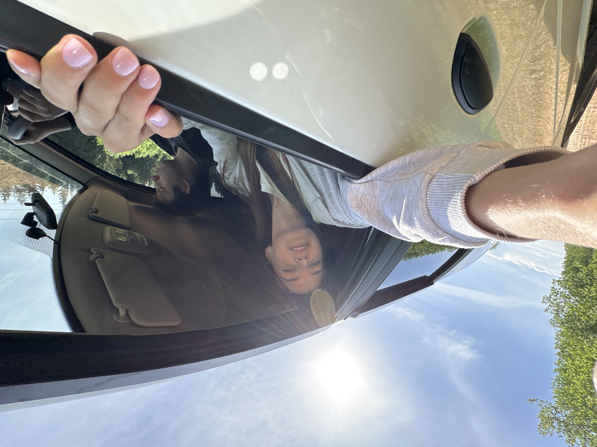 A woman is holding onto the open door frame of a white car, with the interior of the car visible. The sky, with the sun and some clouds, is visible through the open car window. The woman is looking at the camera. Trees are visible in the background. The location cannot be confirmed as Verkhnyaya Khokhlovka, Russia.