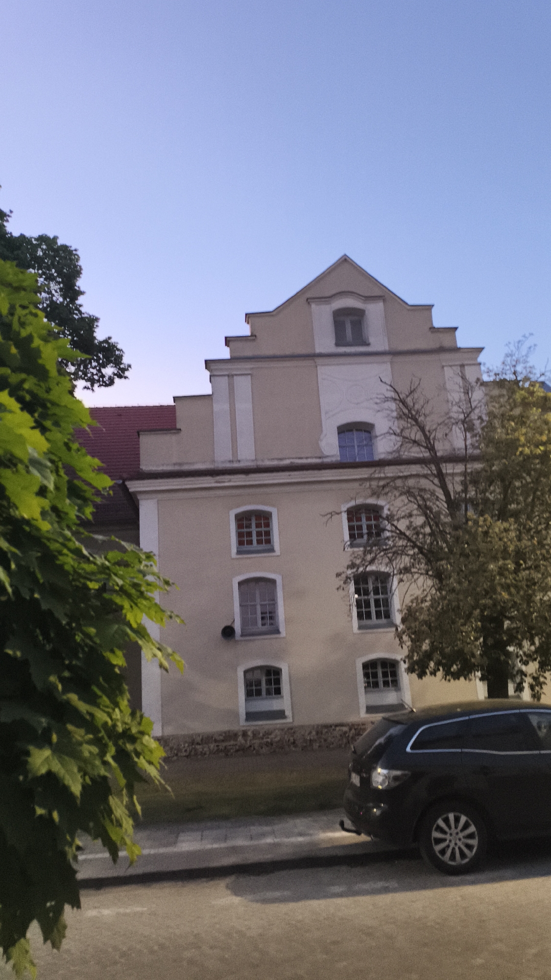 The image depicts a four-story beige building with white trim located in Nowy Tomyśl, Poland. The building features a red roof, symmetrically placed windows, and a decorative gable. To the left of the building, green foliage partially obscures the view. To the right, a tree with sparse leaves stands in front of the building. A black car is parked on the street in front of the building. The sky is a clear gradient of blue, suggesting dusk or dawn. The scene is likely on a street, as evidenced by the pavement in the foreground.