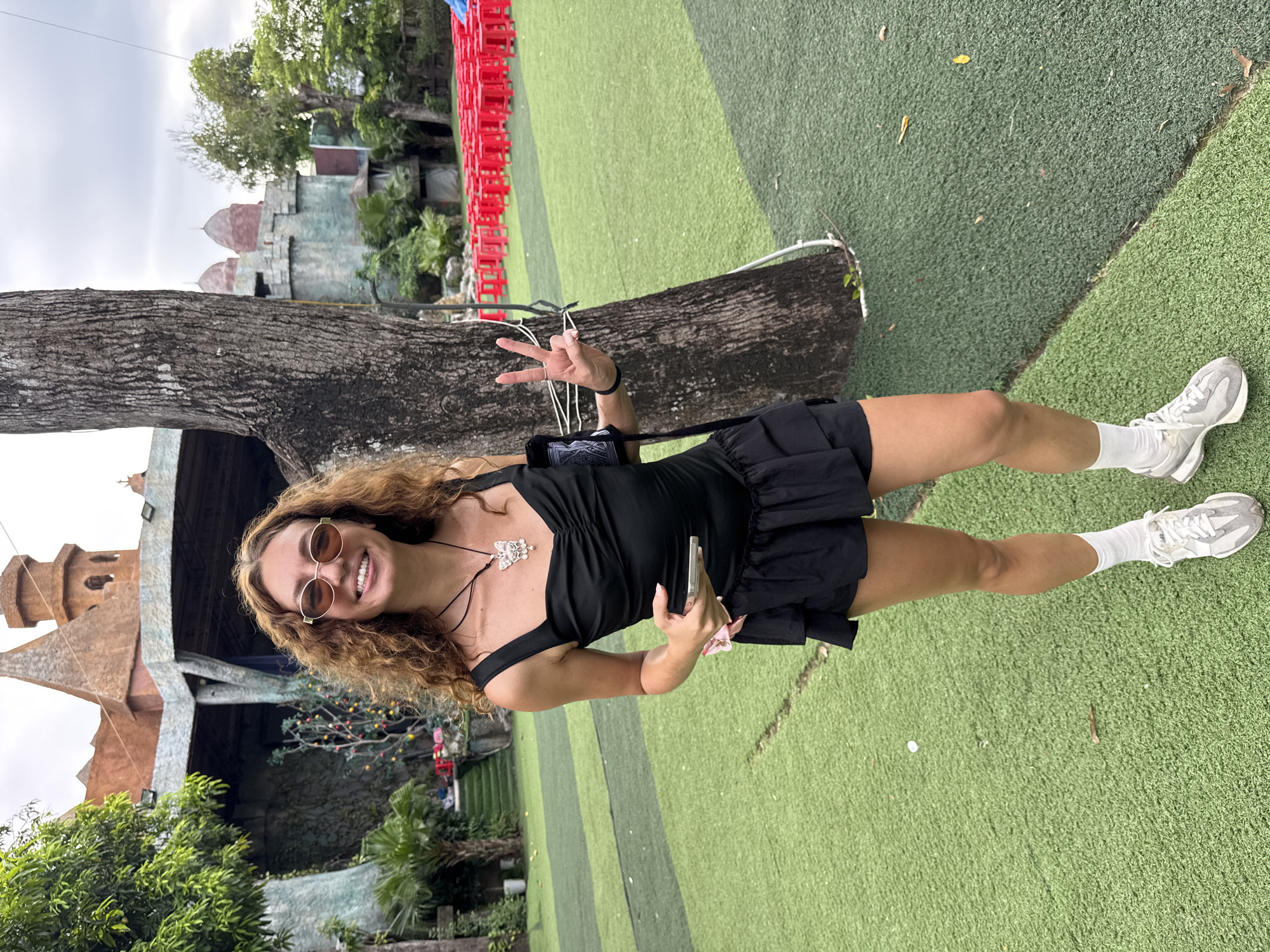 A young woman with curly brown hair and sunglasses stands on an artificial turf field, smiling at the camera and holding up two fingers in a peace sign. She is wearing a black dress with ruffled trim, white socks, and grey sneakers. A large tree trunk is to her right, and in the background, there is a building resembling a castle and rows of red plastic chairs. The environment suggests an outdoor entertainment or event venue.